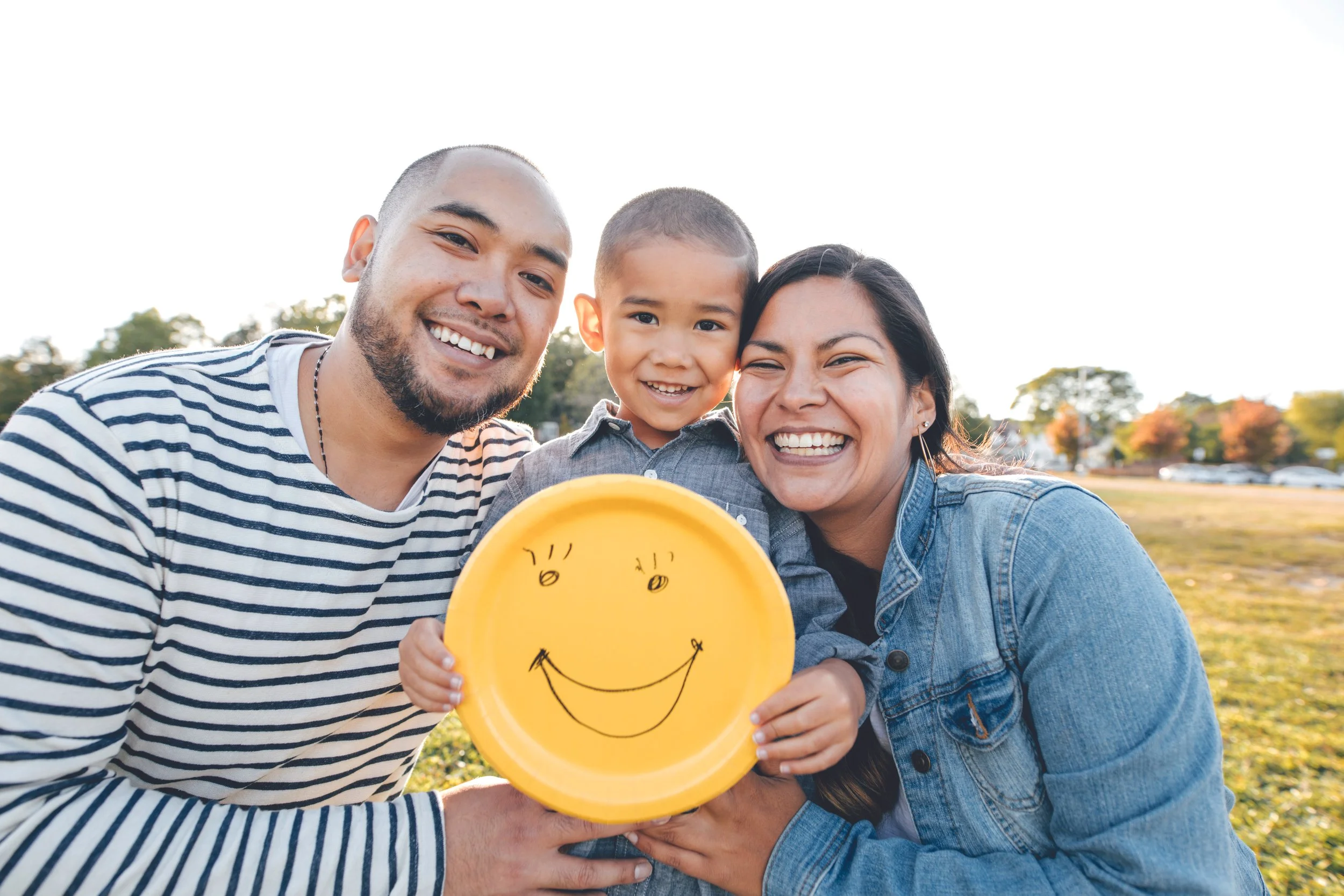 A happy family of three, a man, a woman, and a young boy, taking a selfie outdoors during daytime. The woman and man are smiling, holding a yellow plate with a smiley face drawing, and the family is standing on a grassy field with trees in the background.