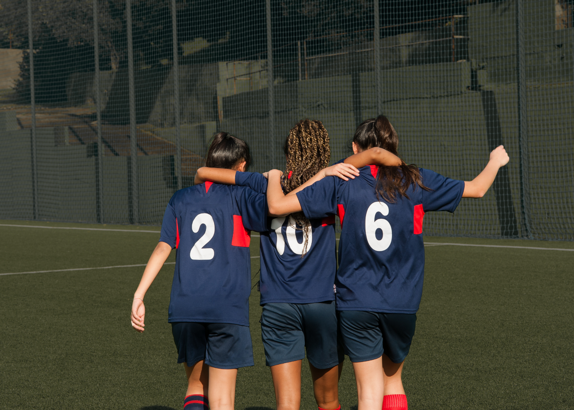 Three young female soccer players in blue uniforms walking arm-in-arm on a soccer field.