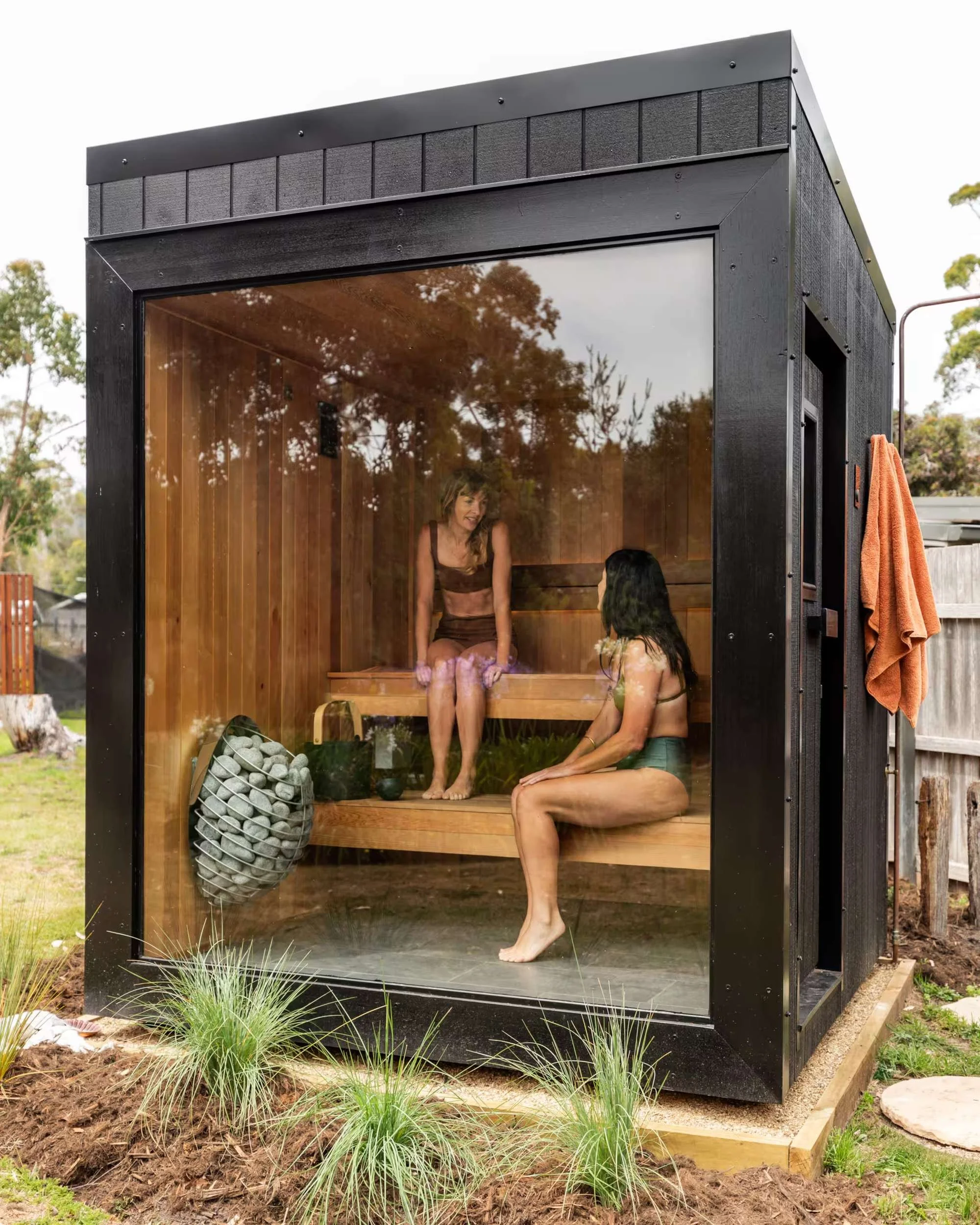 Black hot stone sauna with a glass front, set in the garden with two guests inside