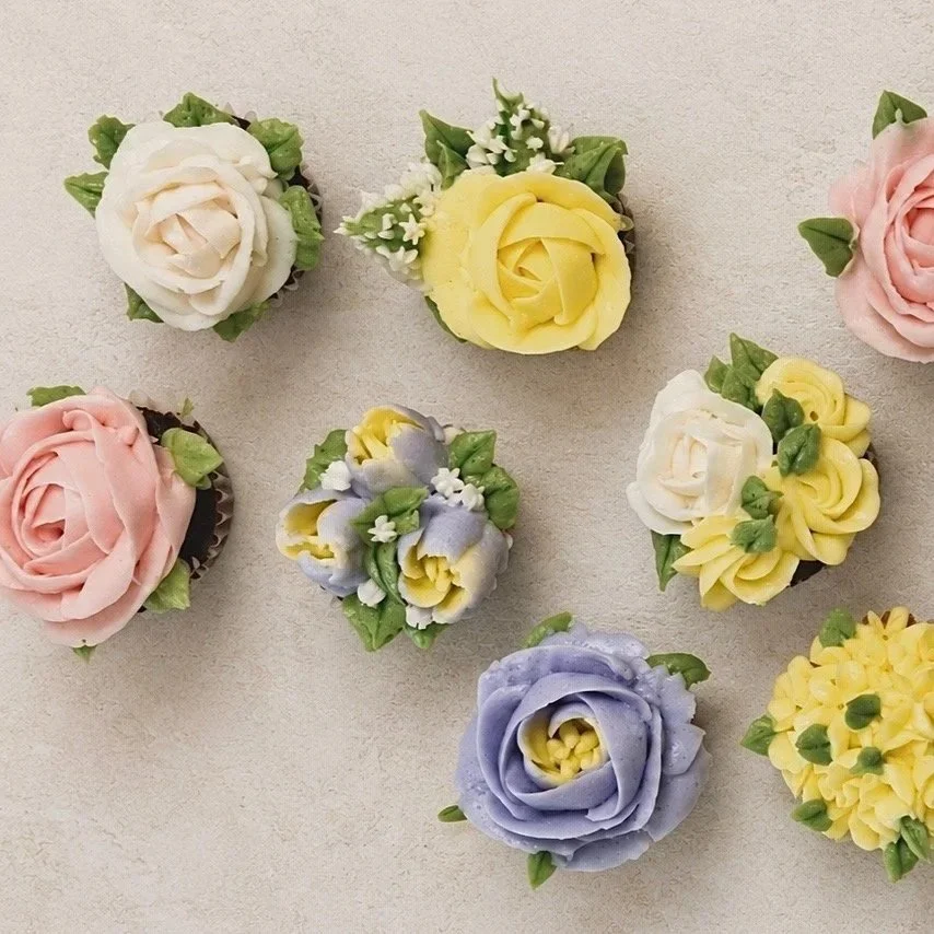 Assorted decorative cupcakes topped with pastel-colored rose and floral designs on a light background.