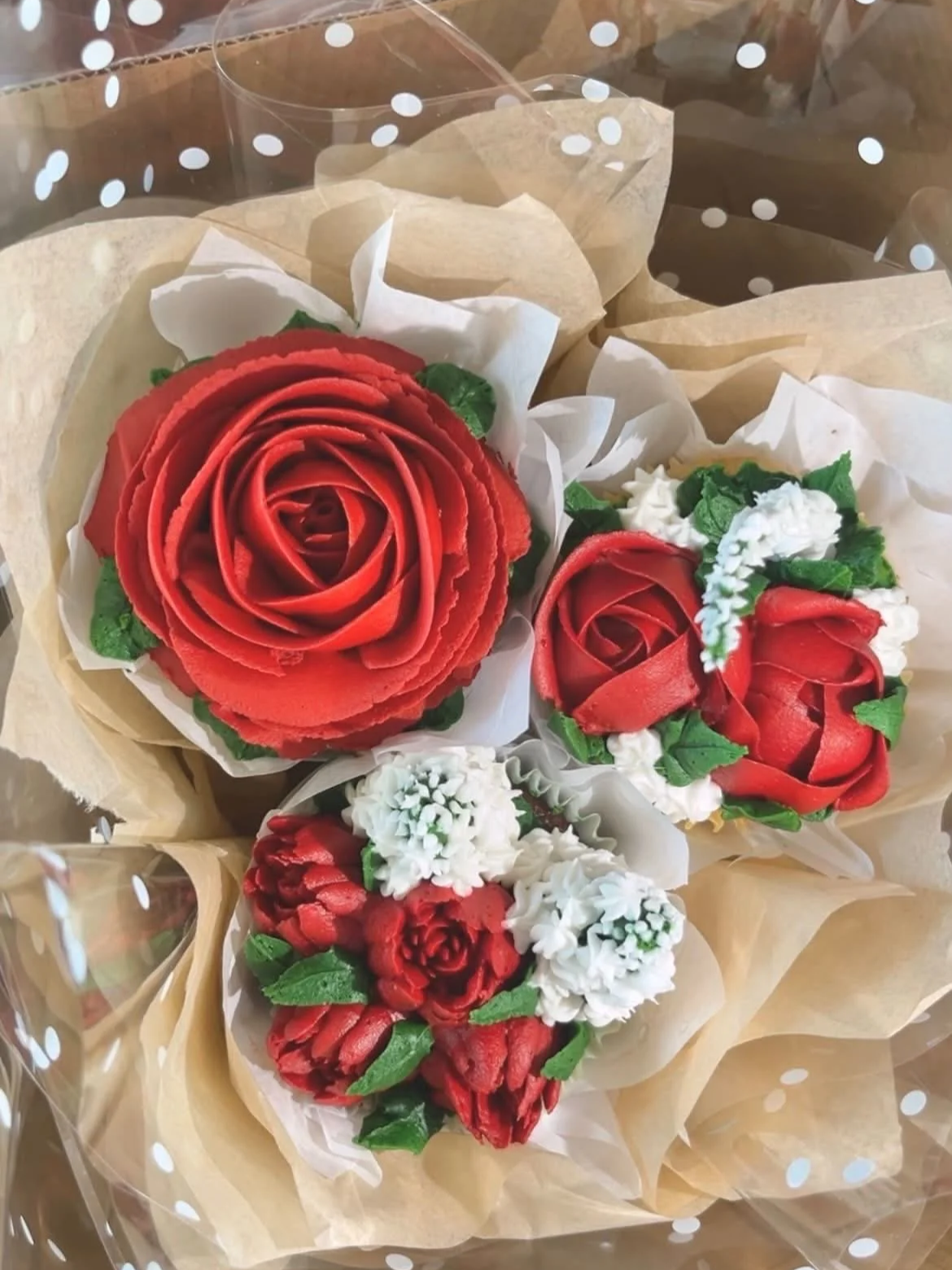 Bouquet of decorative artificial flowers resembling roses and other flowers, wrapped in beige and white paper, inside a clear polka-dotted plastic wrap.