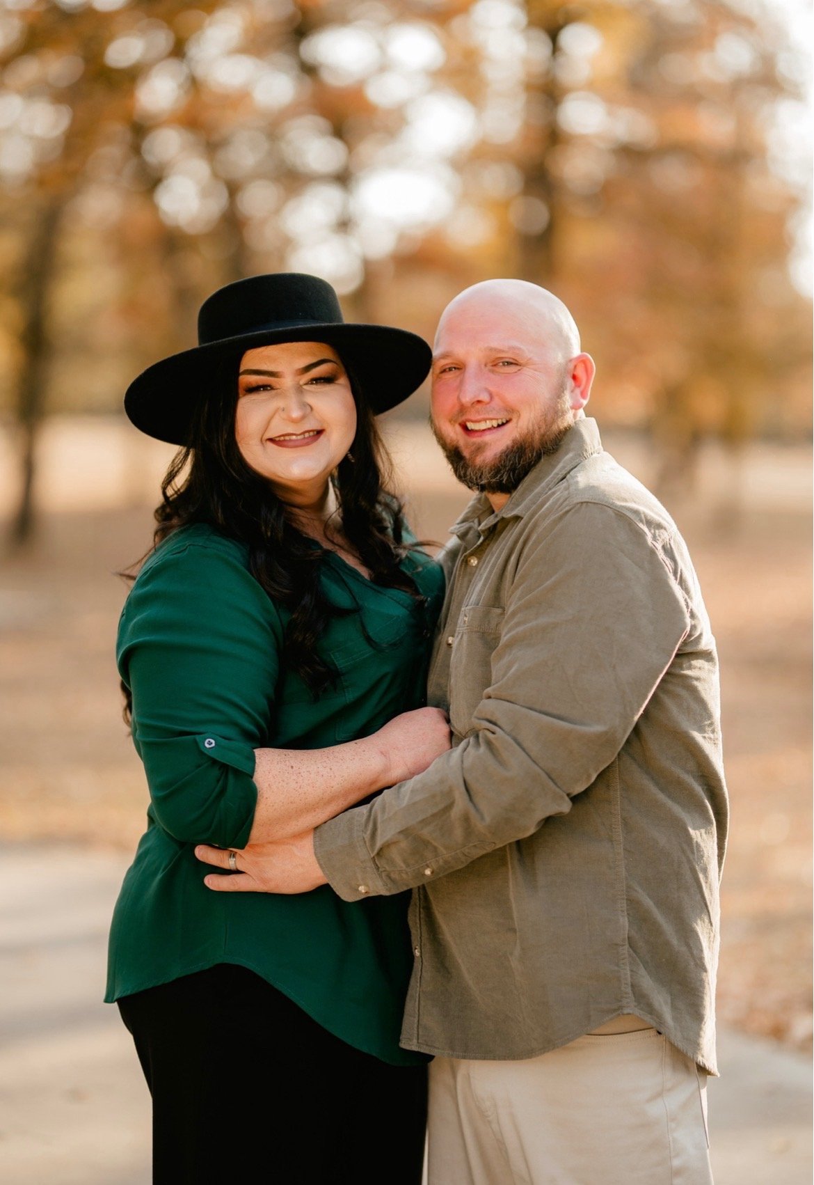 A smiling couple standing closely together outdoors with autumn trees in the background. The woman wears a green dress and a wide-brimmed black hat, and the man wears a beige jacket and has a beard.