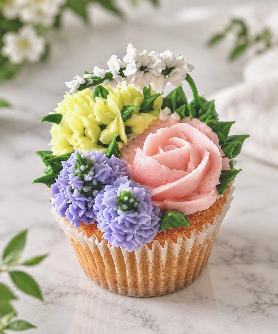 Cupcake decorated with pastel pink frosting shaped like a rose and edible floral decorations in purple, yellow, white, and green.