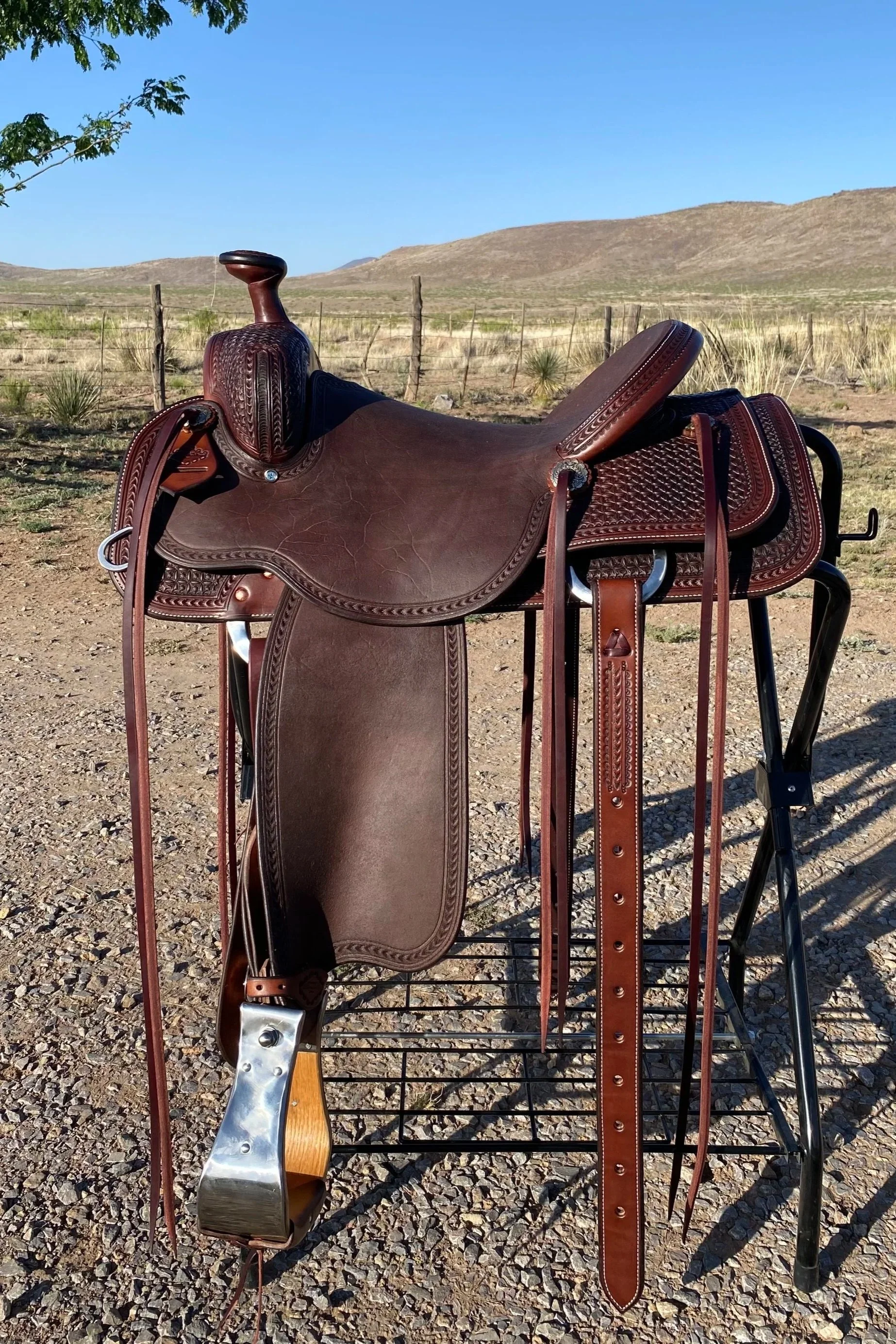 A saddle resting on a black metal saddle stand outdoors in a rural area with dry grass, a wooden fence, and rolling hills in the background under a clear blue sky.