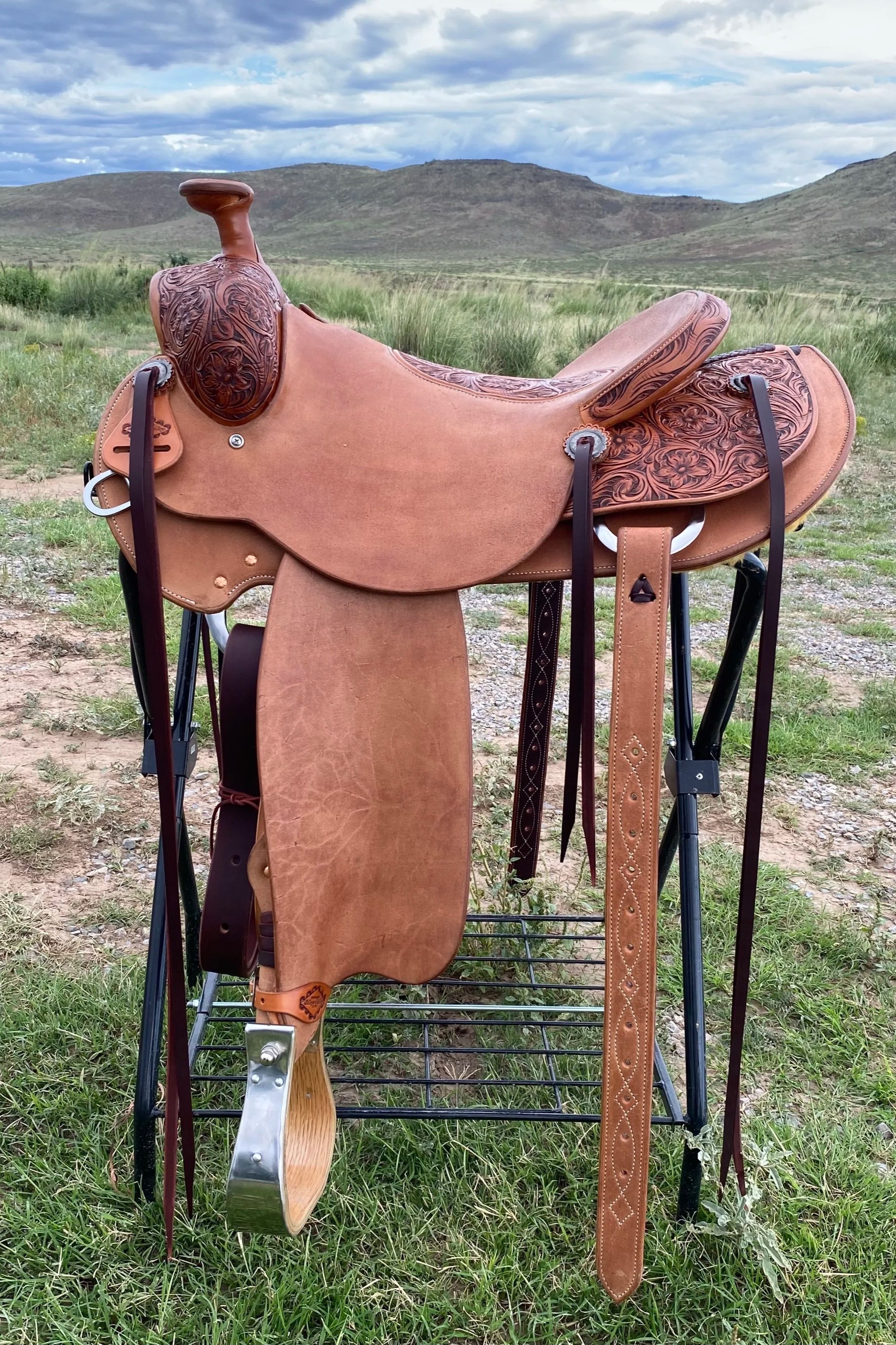 A Western-style saddle with tooled leather details, hanging on a stand outdoors in a grassy field with mountains and cloudy sky in the background.