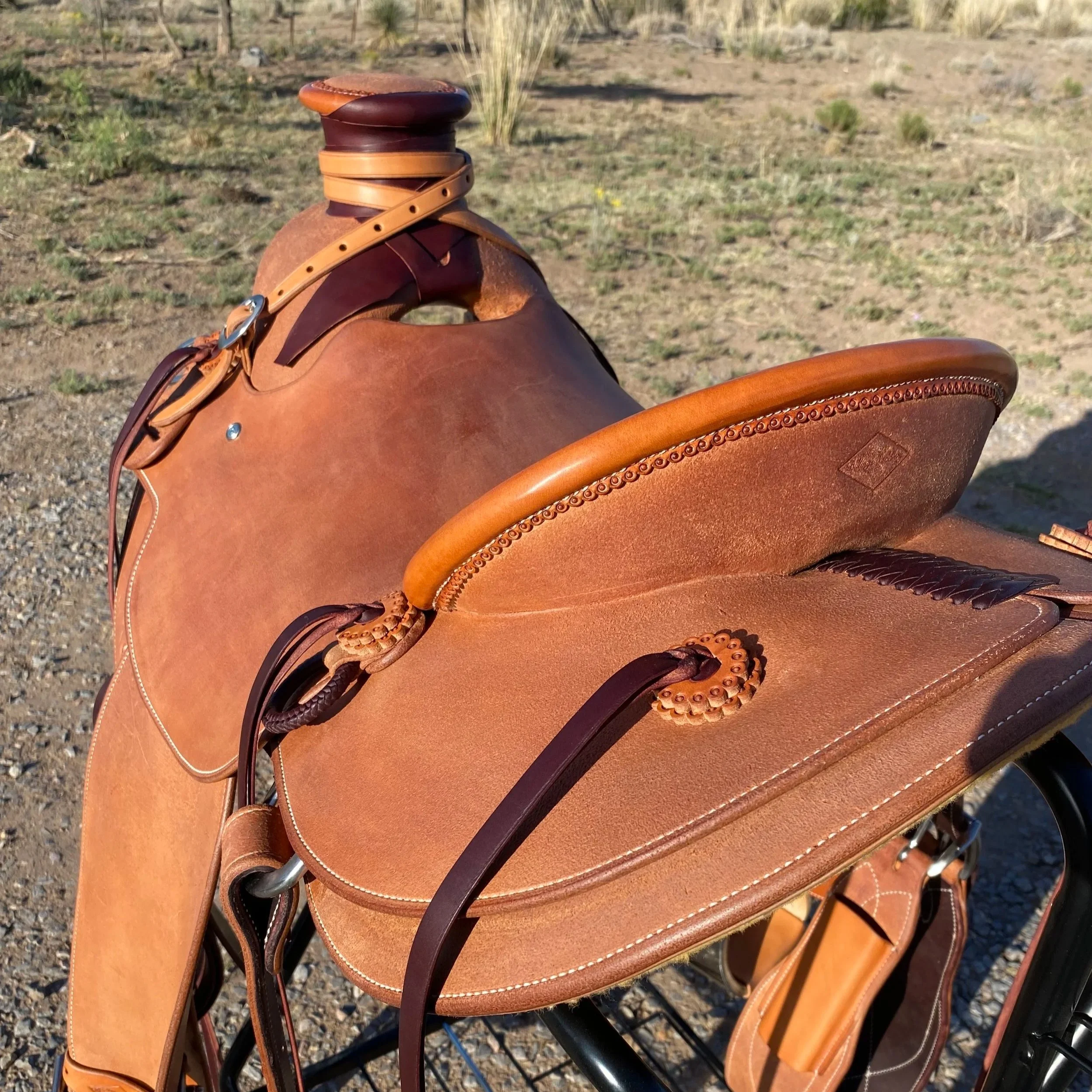 Close-up of a brown leather saddle with decorative stitching, a leather strap, and metal hardware, on a horse outside on a dirt and grass terrain.