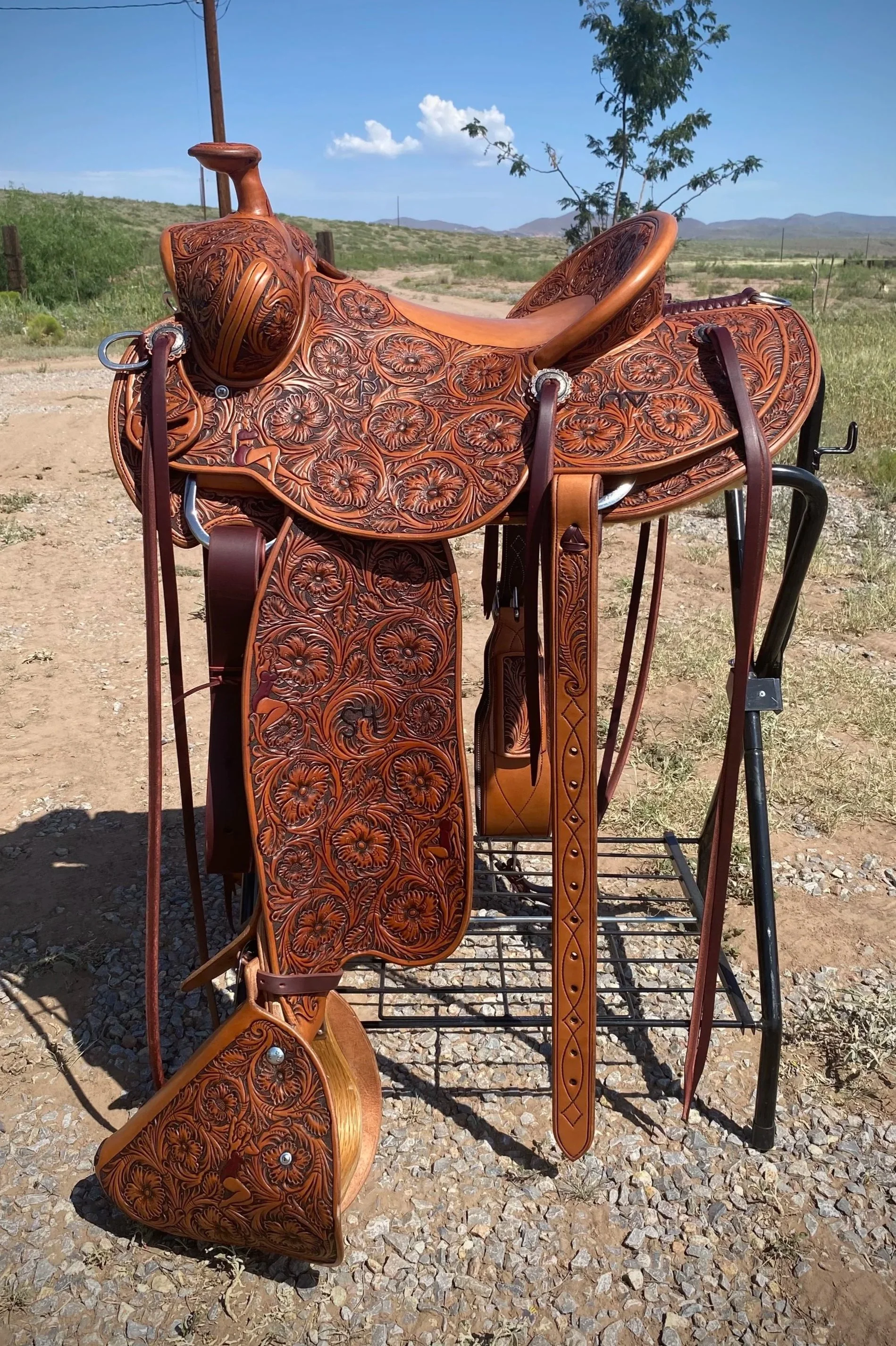 A leather Western-style saddle with floral carvings, placed on a saddle stand outdoors on a gravel surface, with a desert landscape, a tree, and mountains in the background under a blue sky with some clouds.