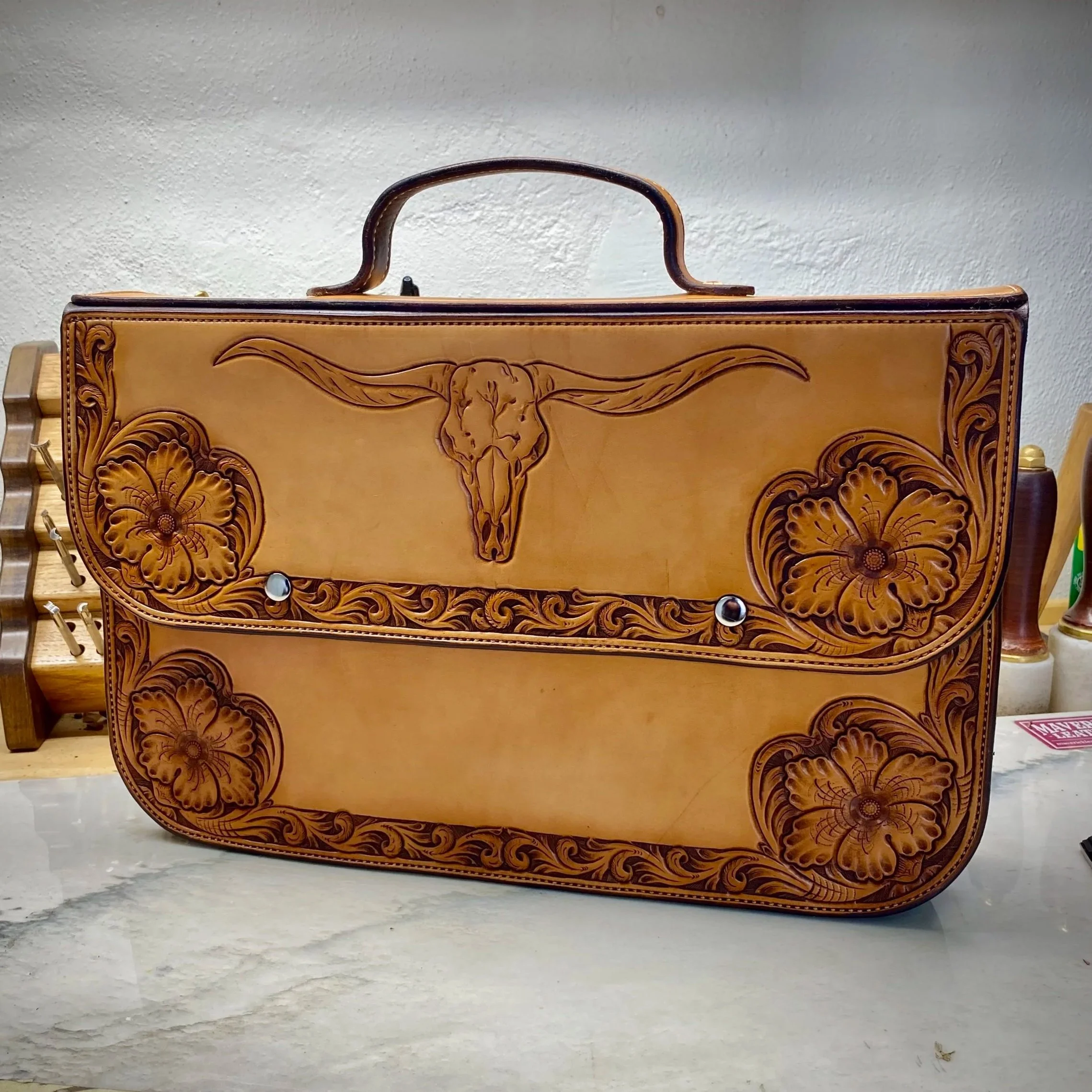 A close-up of a decorative wooden briefcase with carved floral and animal motifs, including a longhorn steer. The box is placed on a concrete surface against a plain white wall.