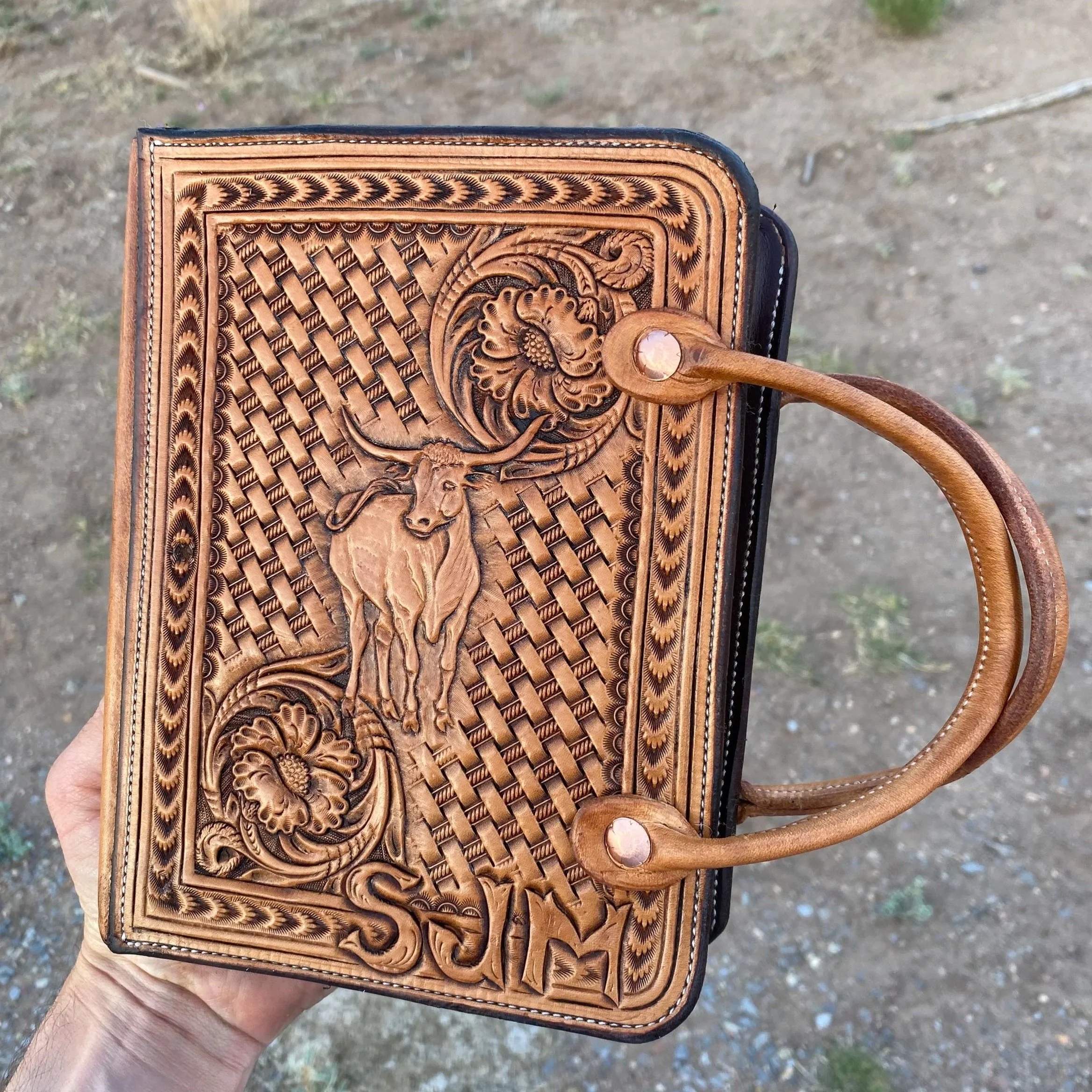 A close-up of a leather book case with intricate carvings, held in a person's hand outdoors with dirt ground in the background.