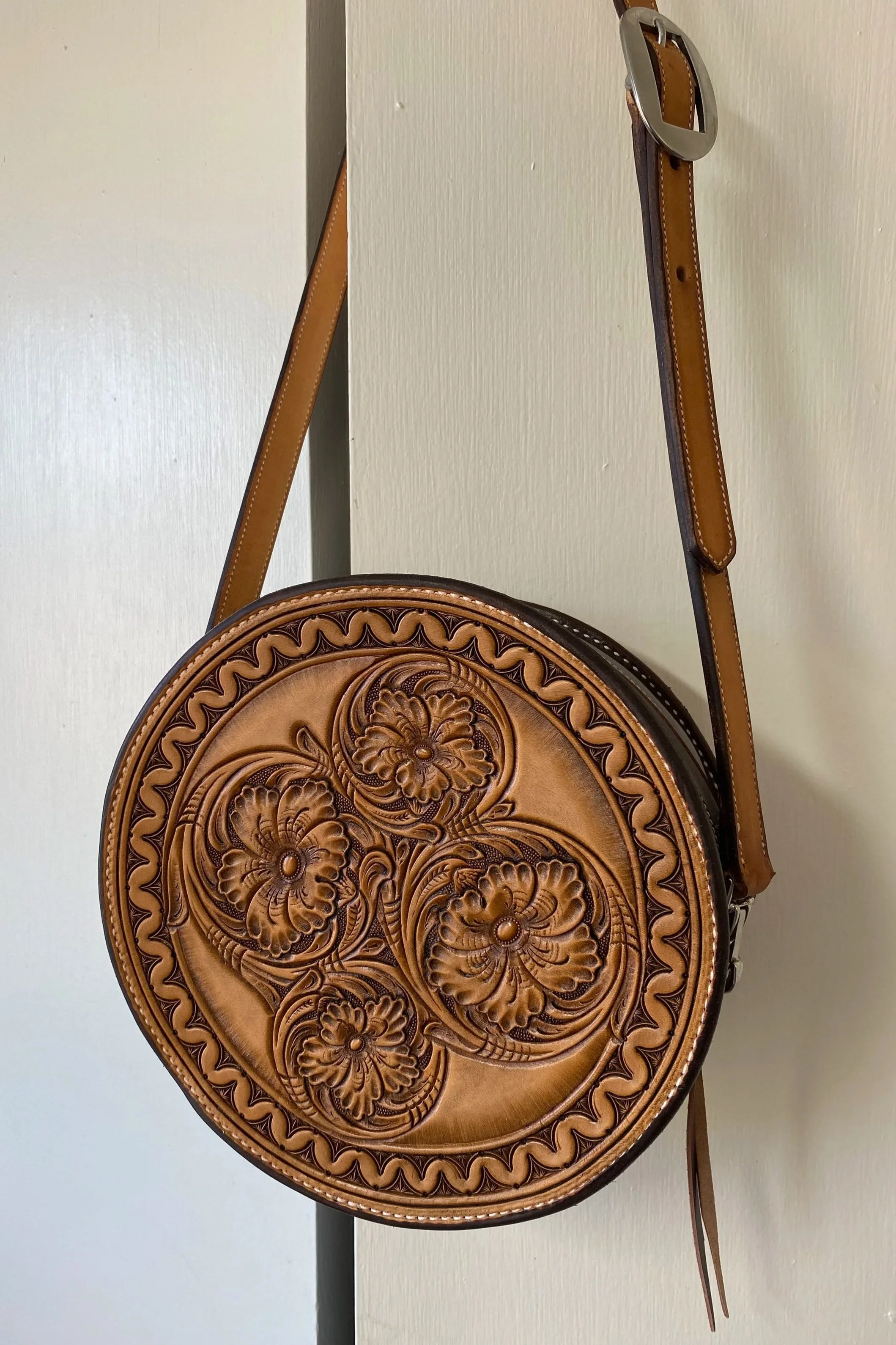 A round, brown leather handbag with intricate floral and leaf carvings on the front, hanging from a strap with a silver buckle on a plain white wall.
