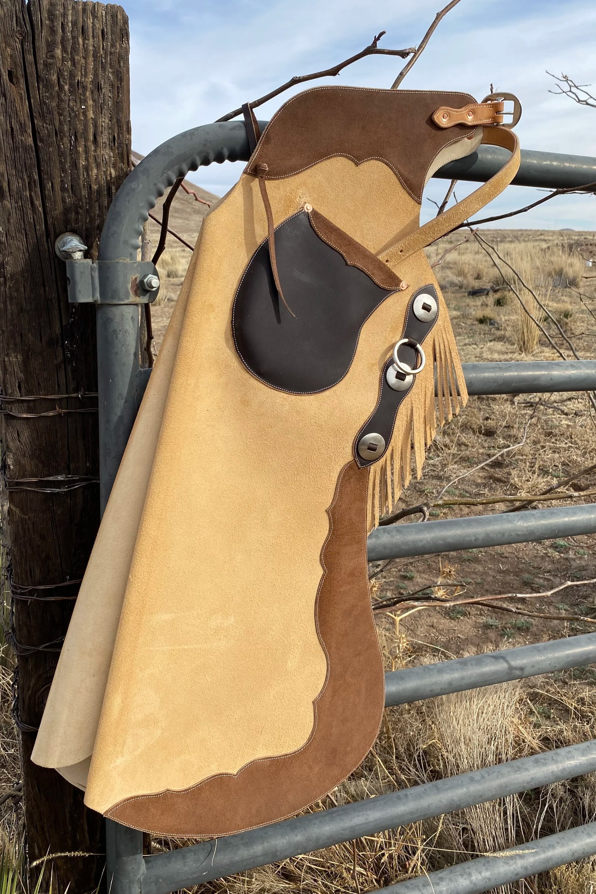 A western-style chaps hanging on a metal gate post in an open desert landscape with dry grass and shrubs, under a cloudy sky.