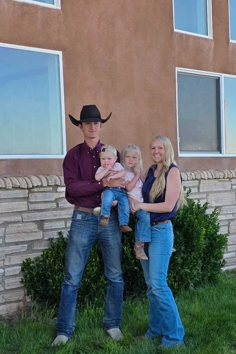 A family of four standing outdoors in front of a building with large windows. The father is wearing a cowboy hat and a maroon shirt, holding a young girl, while the mother stands beside them, smiling, holding a toddler. The family is dressed casually, and there is greenery at their feet.