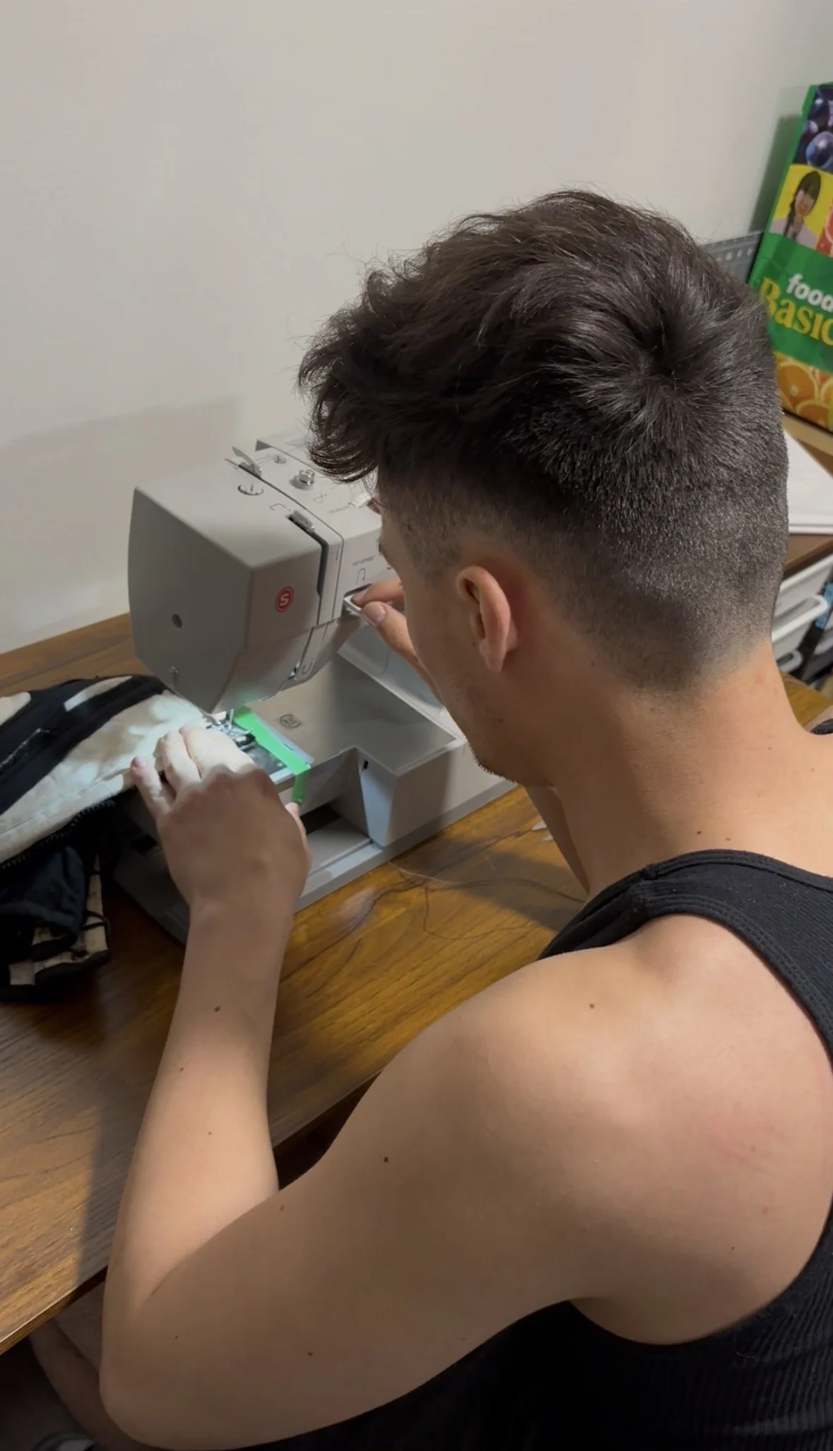 A young man with a fade hairstyle sewing a piece of fabric or clothing on a sewing machine at a wooden table.
