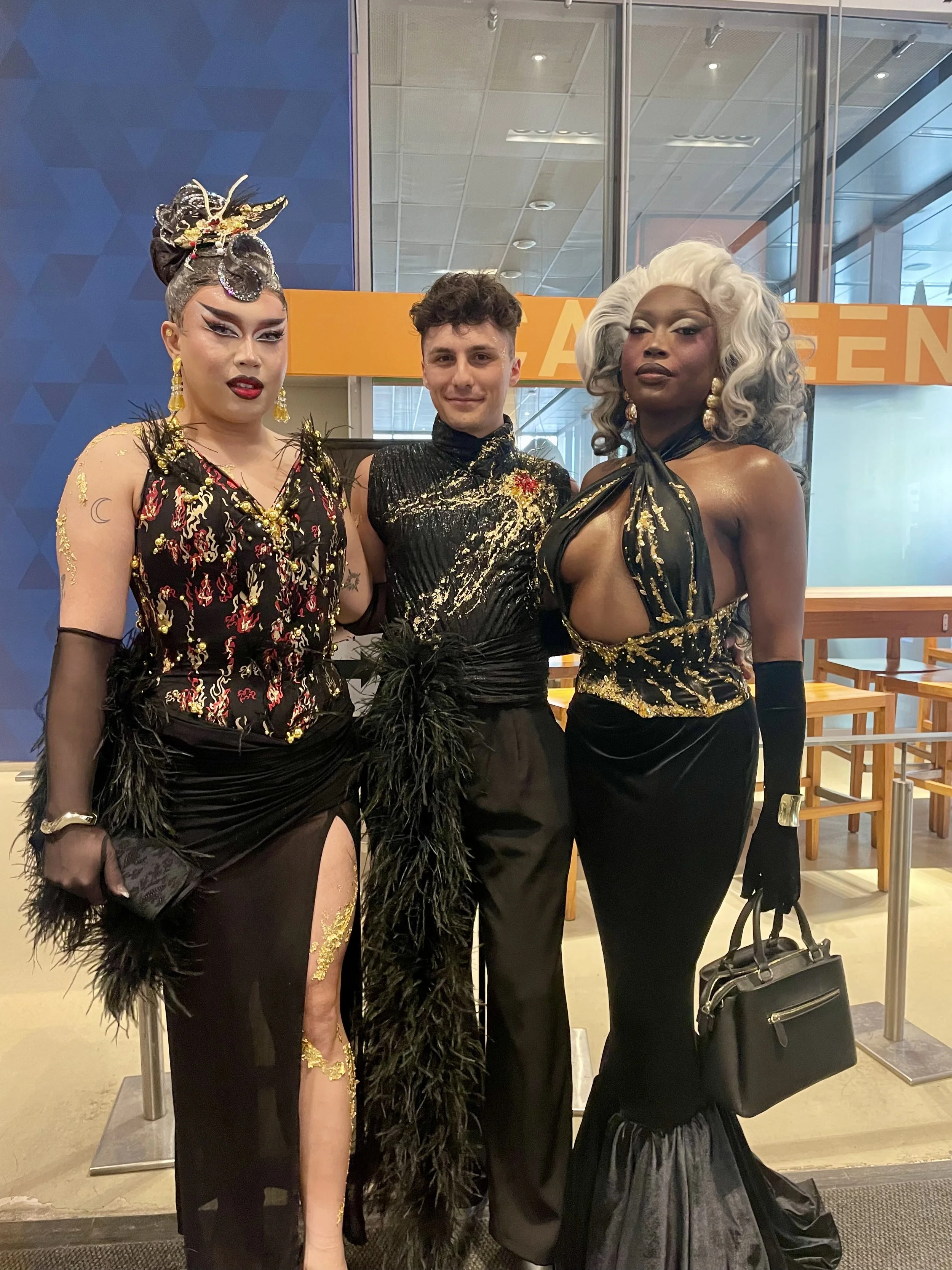 Three individuals in elaborate, glamorous outfits at an indoor event, standing in front of a sign that partially reads 'ATTEN'.