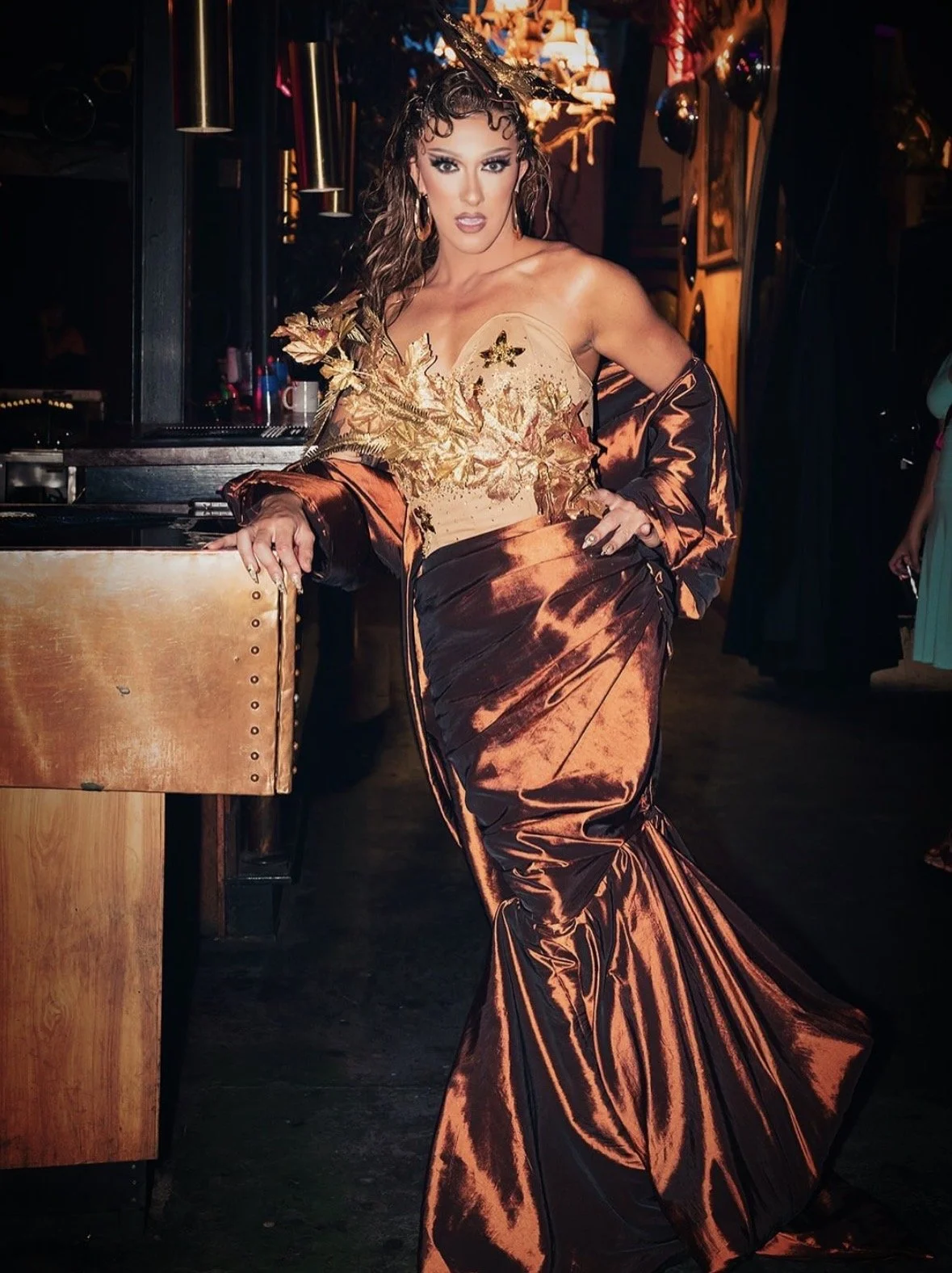 A woman wearing a glamorous, metallic bronze and gold evening gown with a fitted bodice and a long, flowing train. She has curly hair, bold makeup, and is posing in a dimly lit venue with warm lighting and decorative lights in the background.