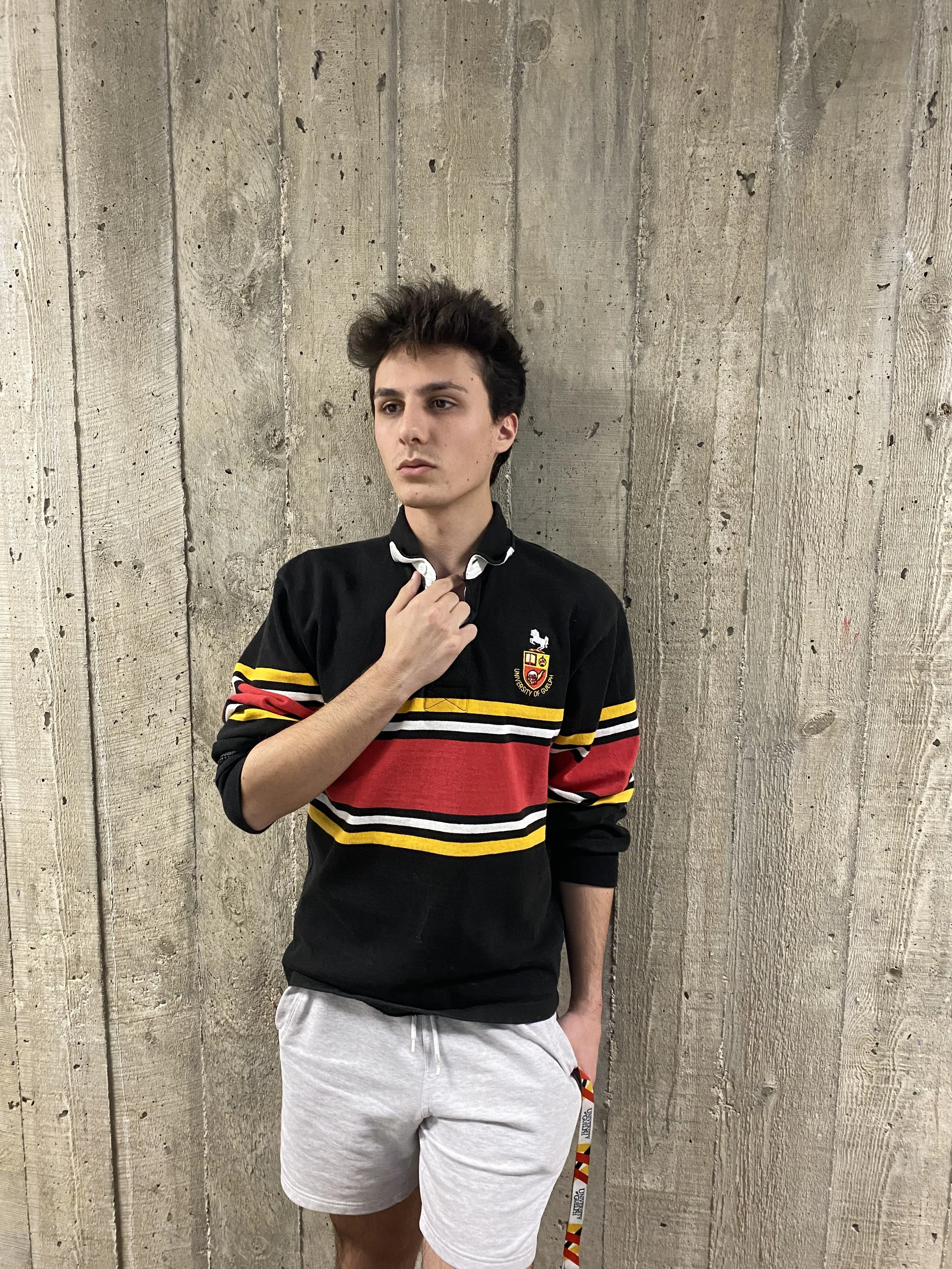 A young man with dark hair and light skin, wearing a black, red, yellow, and white striped rugby shirt with a crest on the chest, and his left hand on his chest, standing in front of a textured concrete wall.