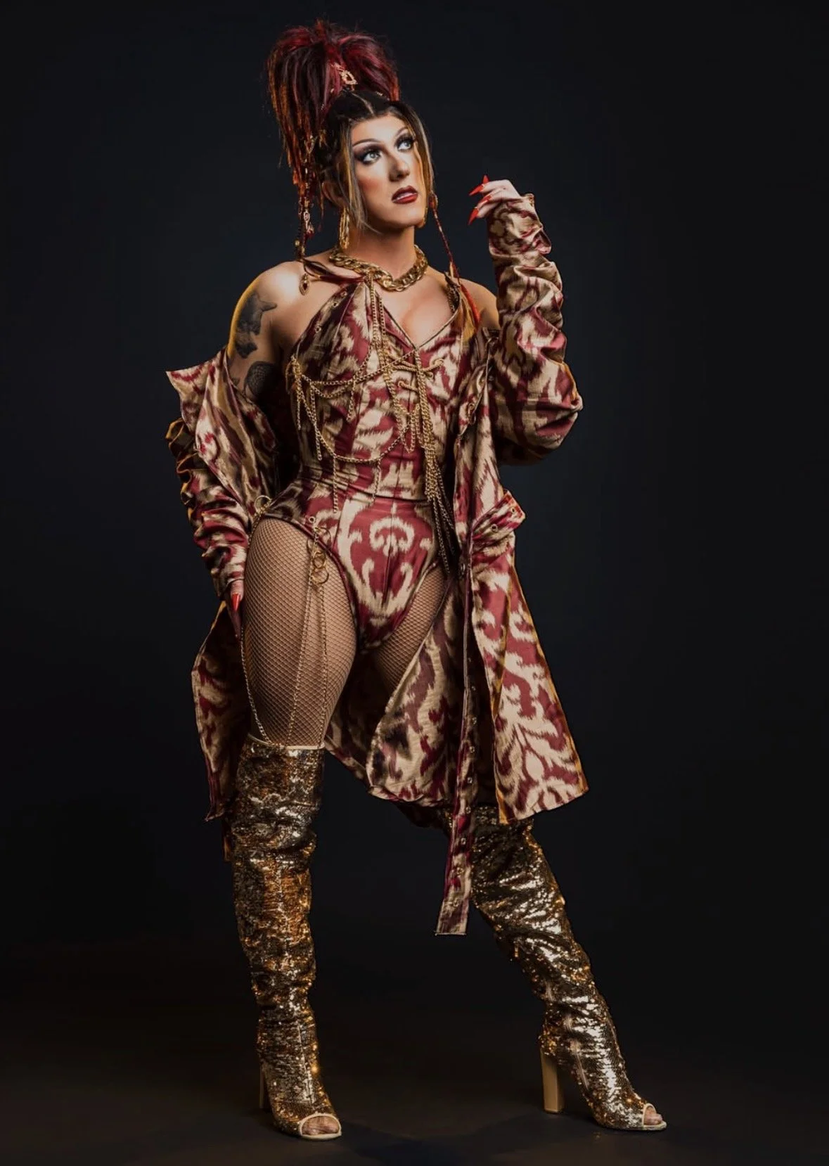 A woman with elaborate hairstyle and makeup wearing a patterned dress, fishnet stockings, and high-heeled boots, posing against a black background.