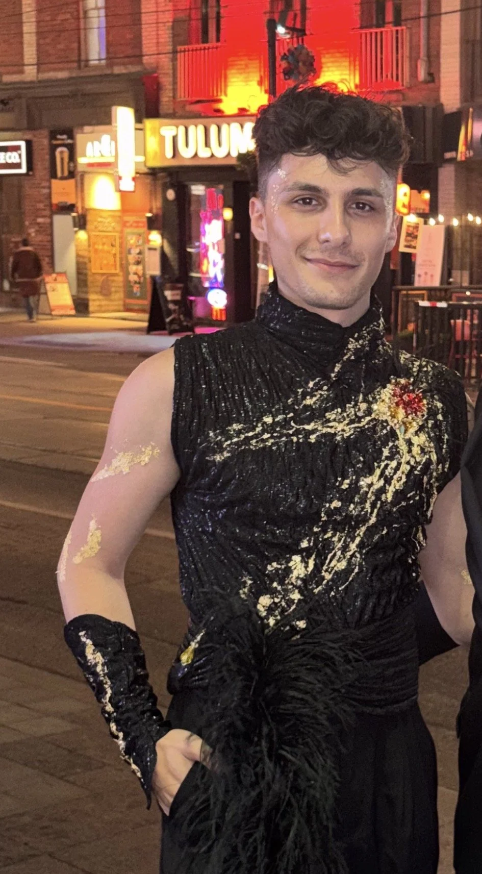 A person standing outside at night, wearing a black sleeveless top with gold paint splatters and a red flower accessory on their chest. They are smiling, with short curly hair and glitter on their face and arm. The background shows a street scene with illuminated signs and buildings.