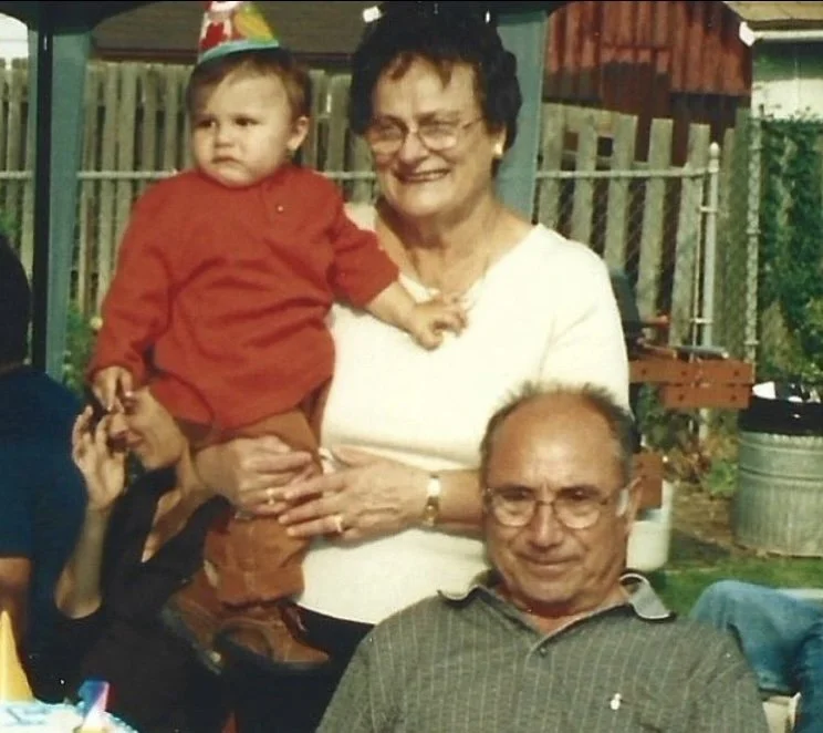 Three generations of a family celebrating outdoors, with a woman holding a young child on her lap, and an older man sitting in front. The background shows a fenced yard with plants and a small shed.