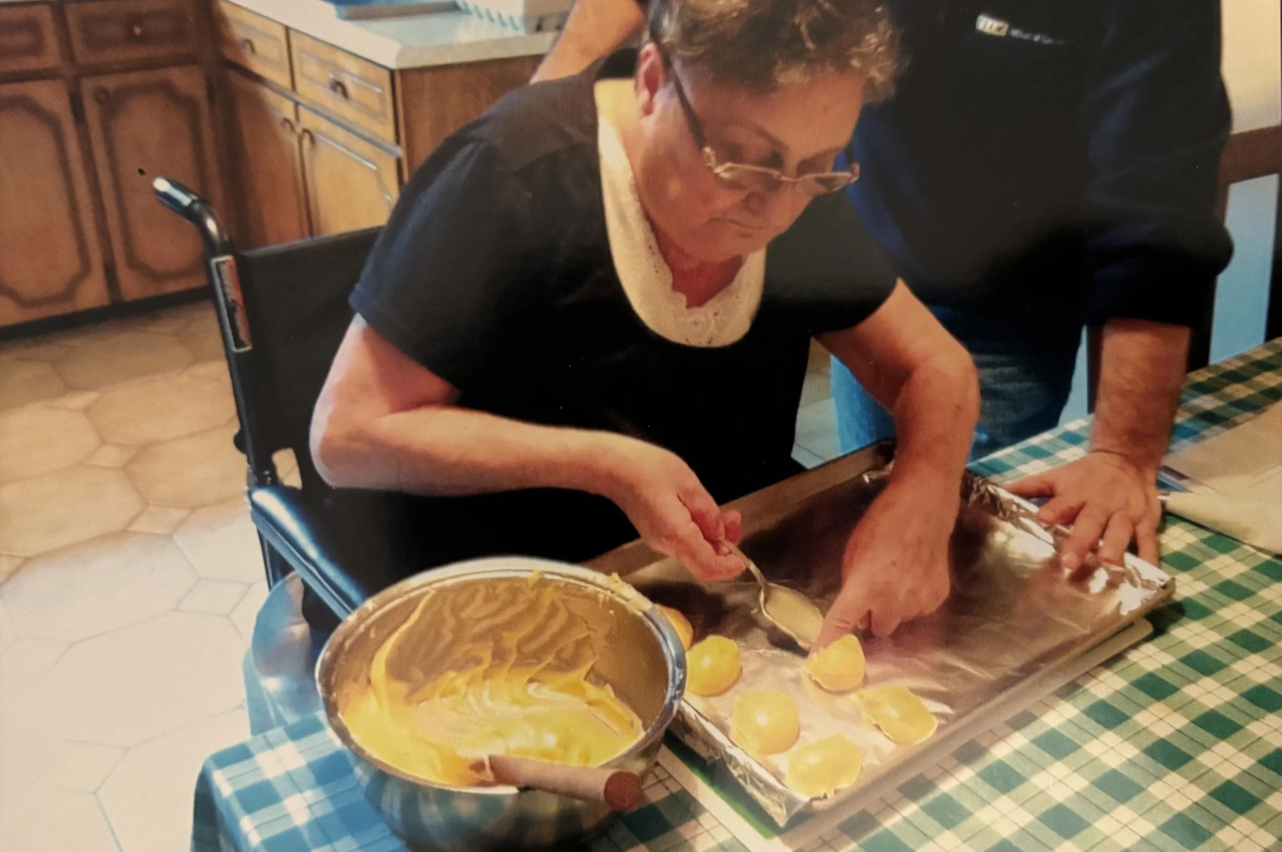 An elderly woman sitting at a kitchen table, filling peeled potatoes into a bowl. A pan of yellow mashed potatoes and a bowl of mashed potatoes are on the table, which has a blue checkered tablecloth.