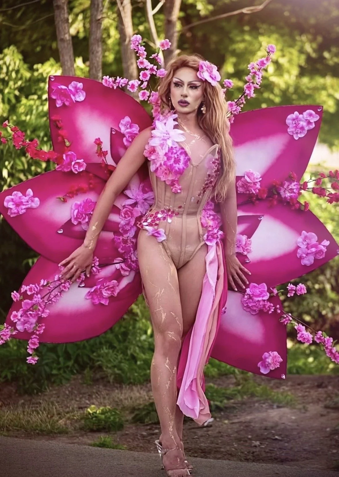 A woman dressed as a fairy with large pink wings, surrounded by pink flowers, standing outdoors in a forested area.