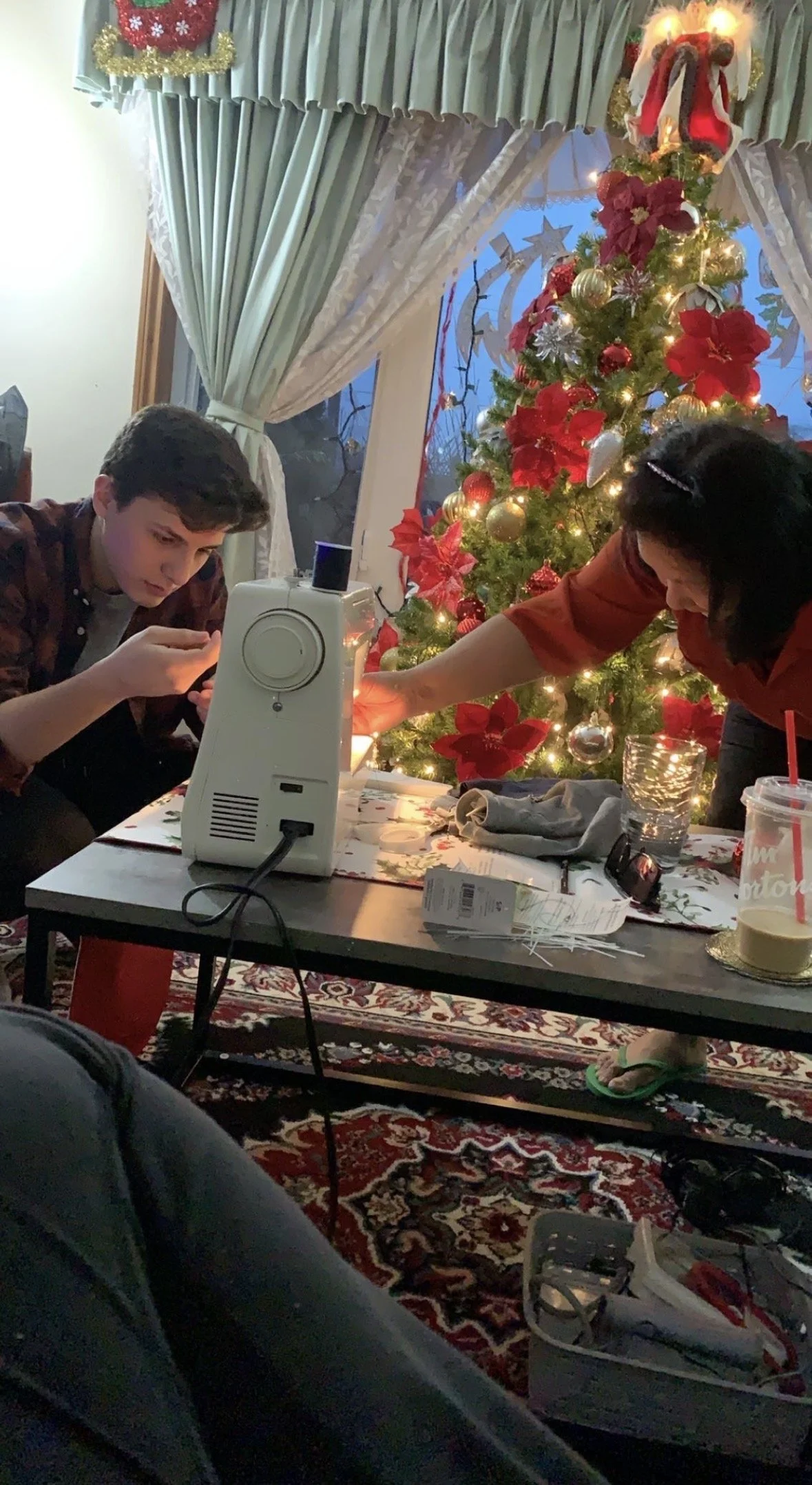 A family gathers around a decorated Christmas tree in a living room, working on a holiday craft or decoration at a table. The room features festive decorations, including curtains and a holiday arrangement with red and gold accents.