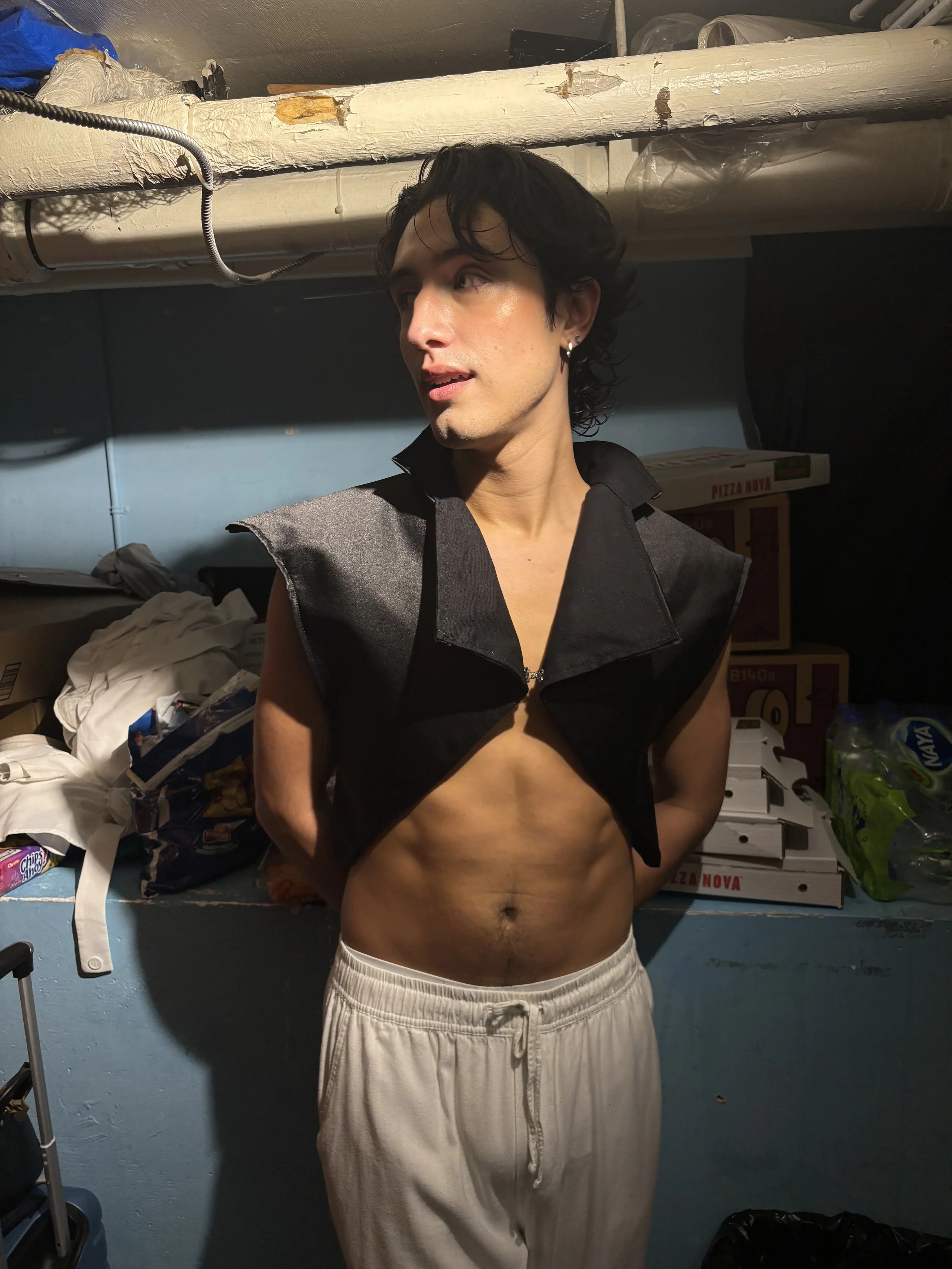 A young man with black curly hair and earrings, shirtless with a black vest and white sweatpants, standing in a cluttered room with boxes, bags, and miscellaneous items around him.