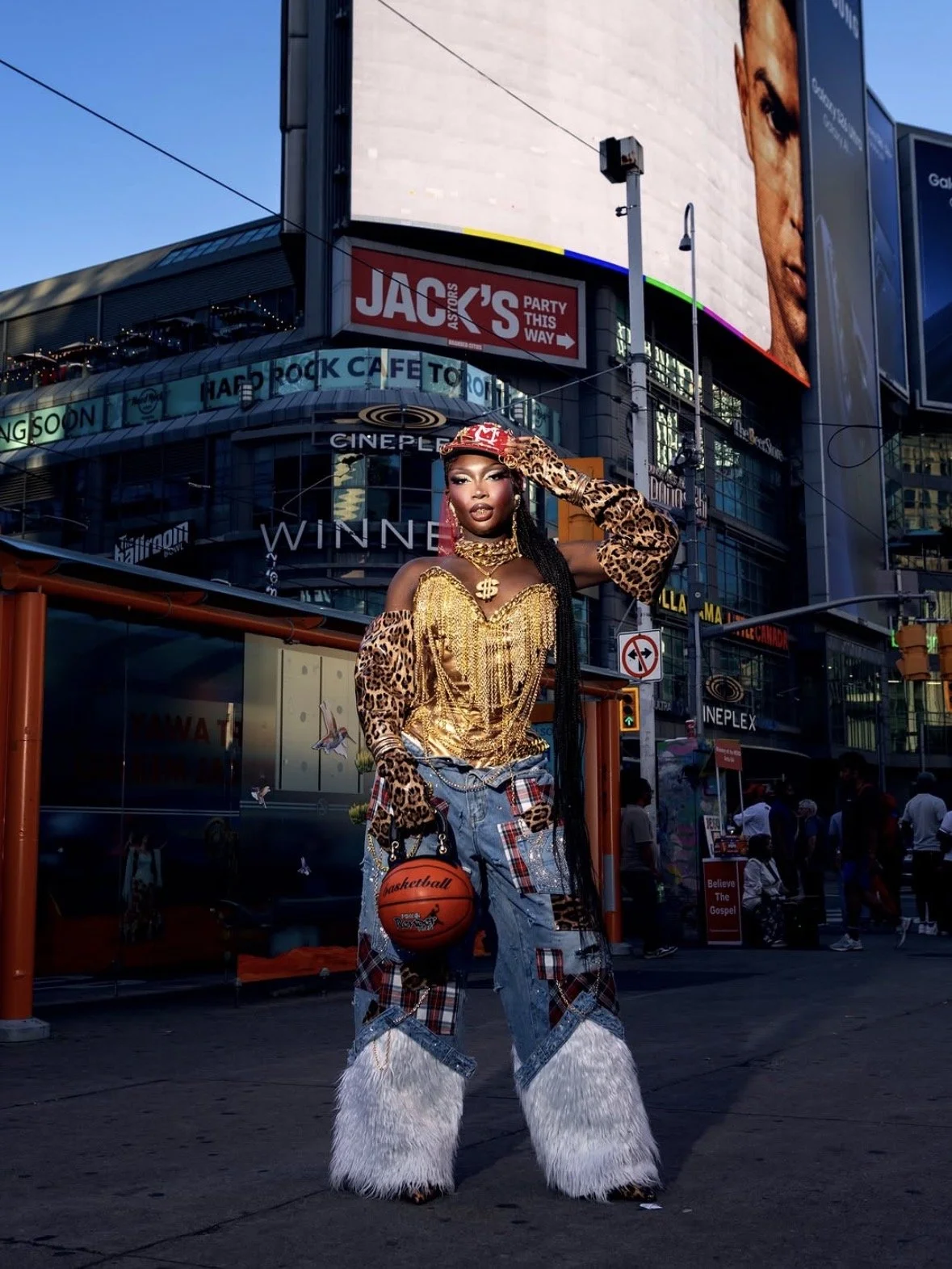 A person with long hair styled in braids, wearing a leopard print hat, gold chain necklace, and a gold top with fringe, dressed in patchwork jeans with furry leg coverings, holding a basketball, standing in a city square with digital billboards and onlookers in the background.