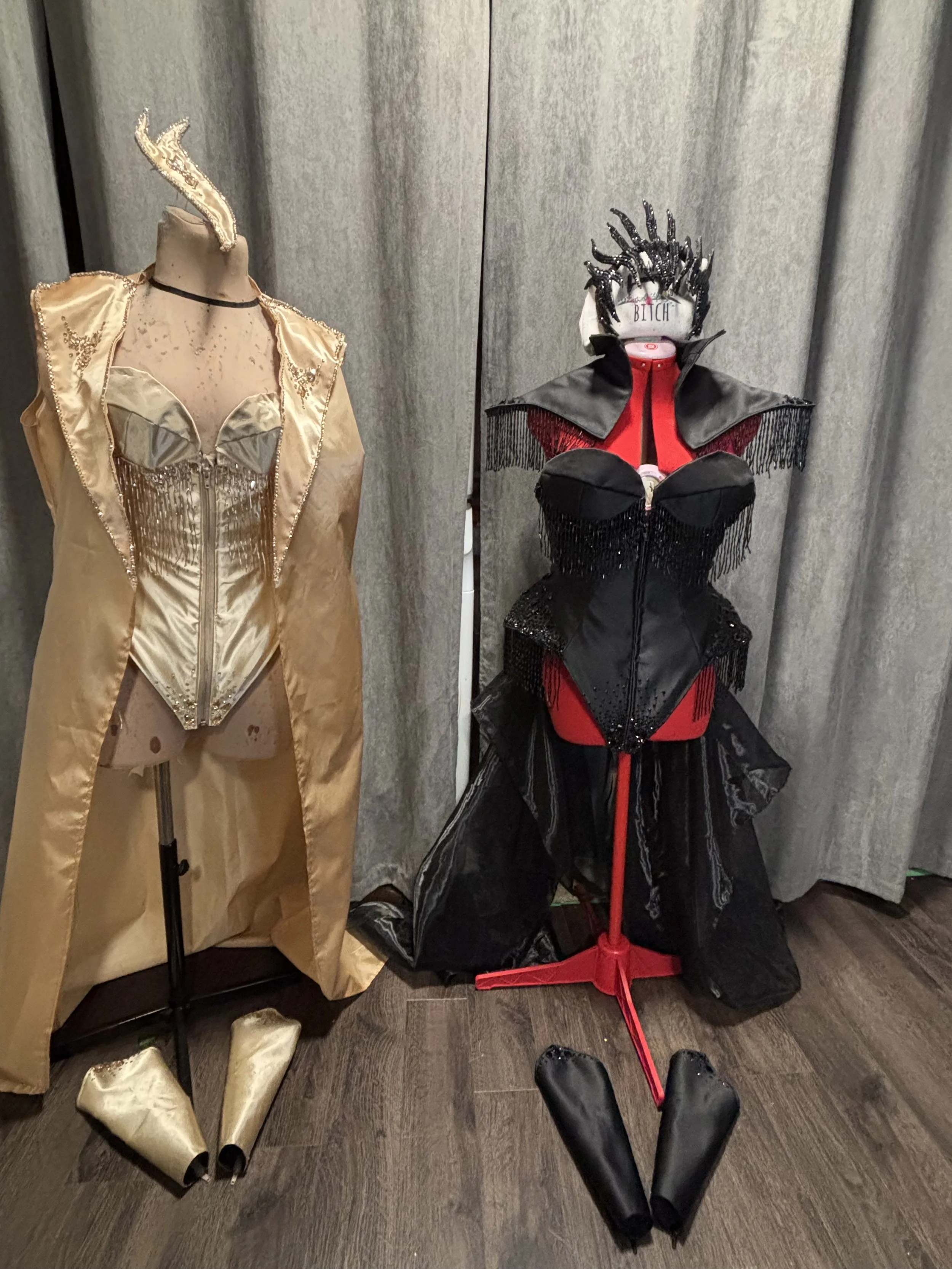 Two mannequins dressed in elaborate costumes and accessories, with one in gold and the other in black and red, positioned against a curtain backdrop.