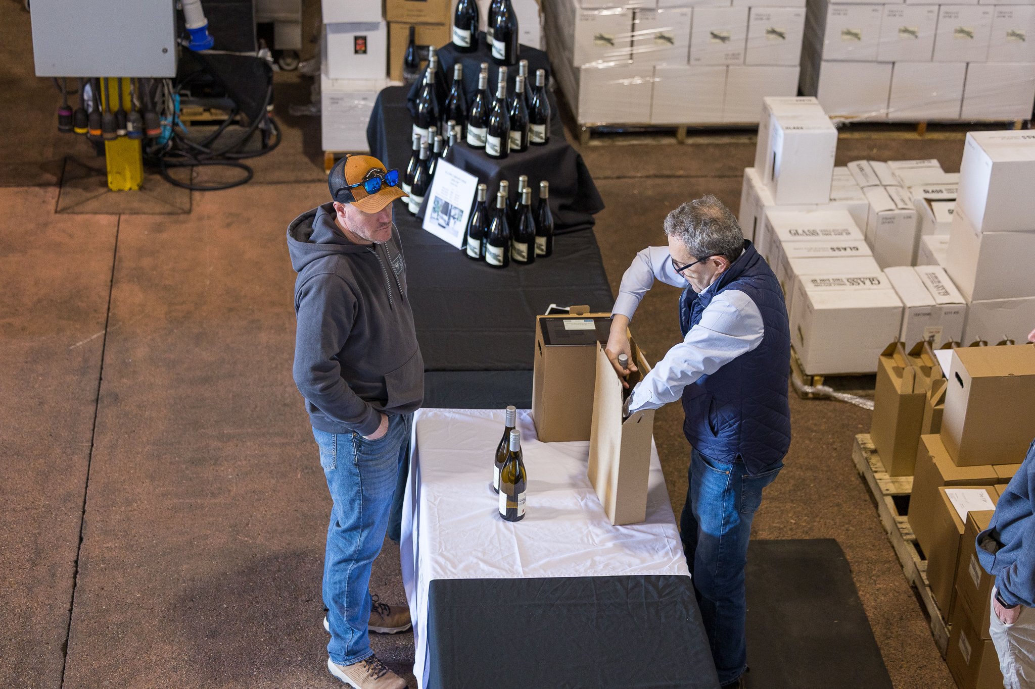 Two men are standing at a table with bottles of wine, with one man placing a bottle into a cardboard box, while the other watches. There are more bottles of wine on the table and shipping boxes behind them, in what appears to be a warehouse or storage facility.