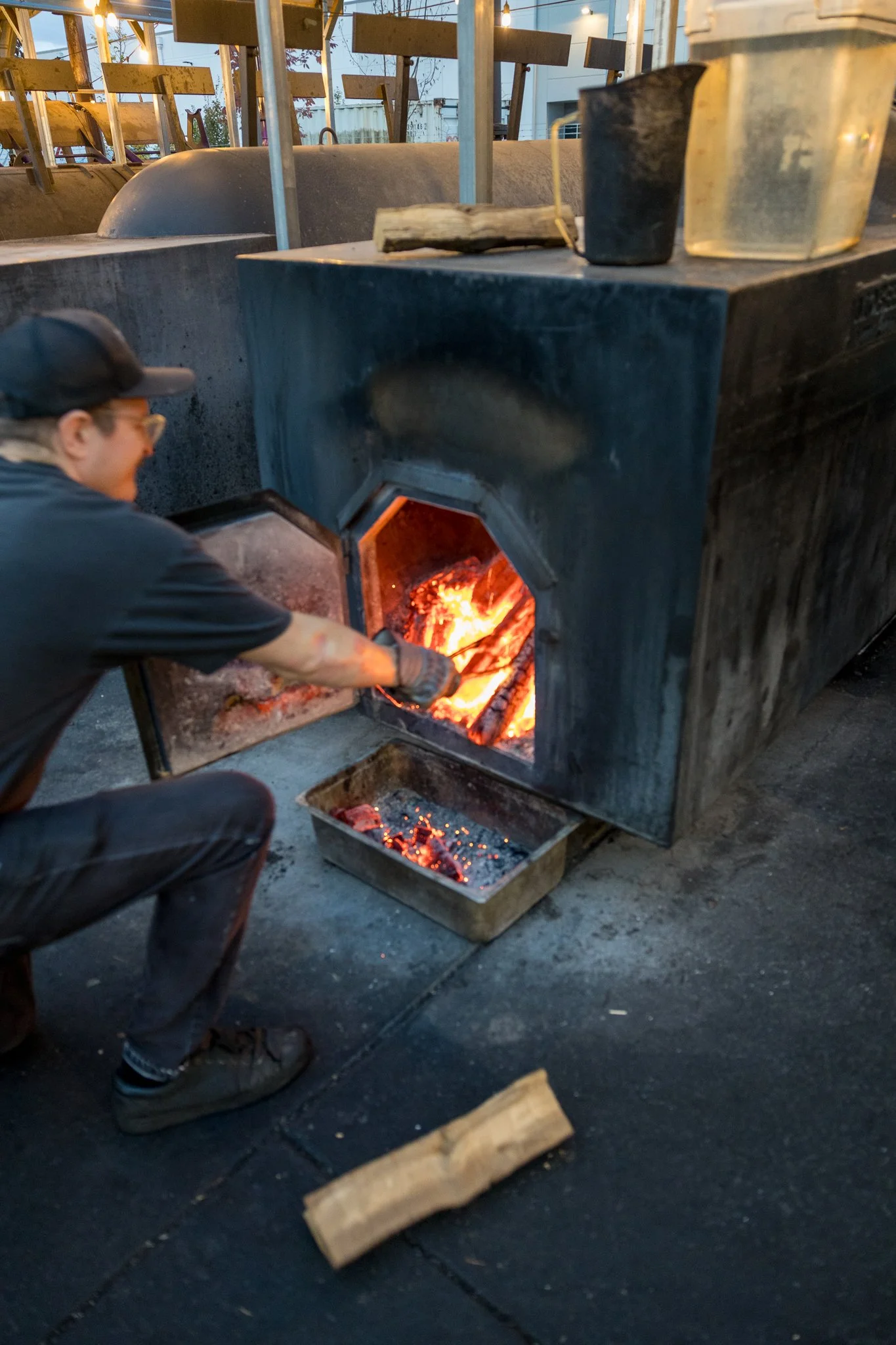 Person wearing glasses and a cap tending to a wood fire inside a large black metal oven, with fire visible through the open door. The outdoor setting includes wooden signs or boards on a fence in the background, and the ground appears to be paved.