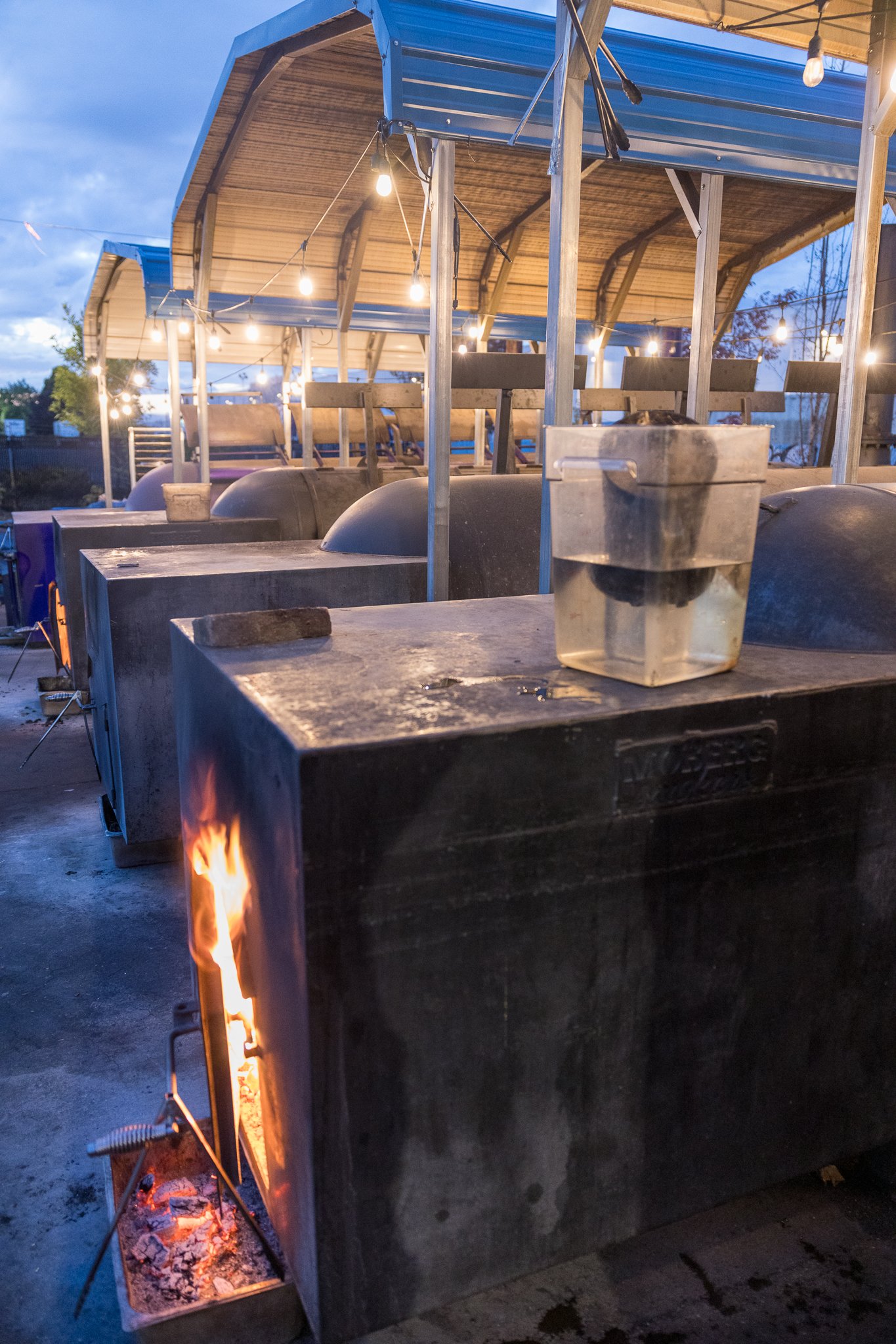 Outdoor barbecue smoker with a fire burning inside, metal construction, with hanging string lights, set against evening sky.