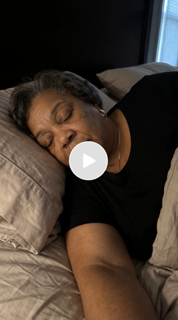 A woman sleeping peacefully in bed with her head on a pillow, wearing a black shirt, near a window with blinds.