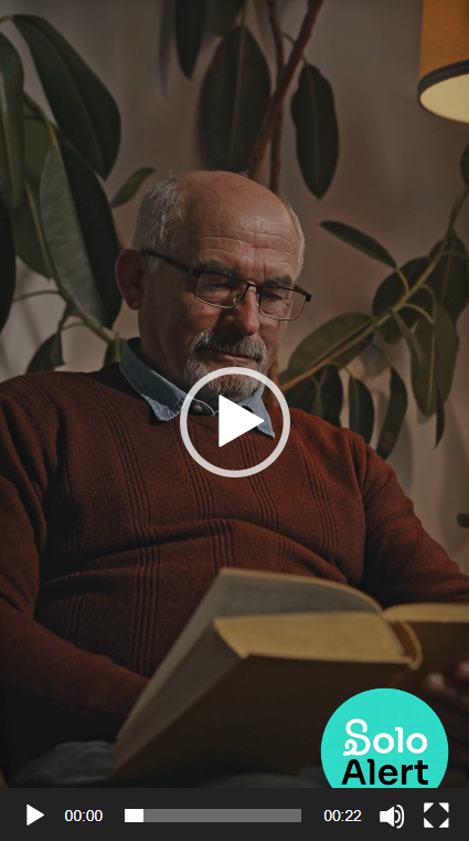 An elderly man with glasses reading a book indoors, with large leafy plants and a lamp in the background.