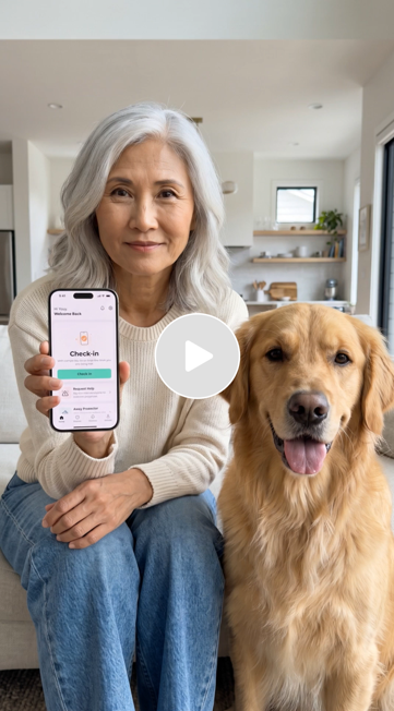 An older woman with gray hair holding a smartphone displaying a check-in app, sitting next to a golden retriever in a bright, modern kitchen.