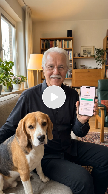 An elderly man with glasses and a gray mustache sitting on a sofa, holding a smartphone displaying a checked-in app, with a beagle dog sitting beside him, in a cozy living room with bookshelves, a lamp, and houseplants.