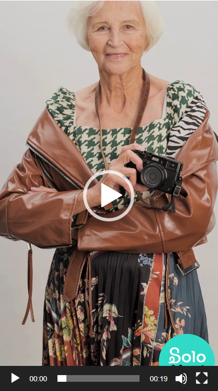 An elderly woman with white hair smiling, holding a camera, wearing a patterned top, a brown leather jacket, and a floral skirt, standing against a plain background.