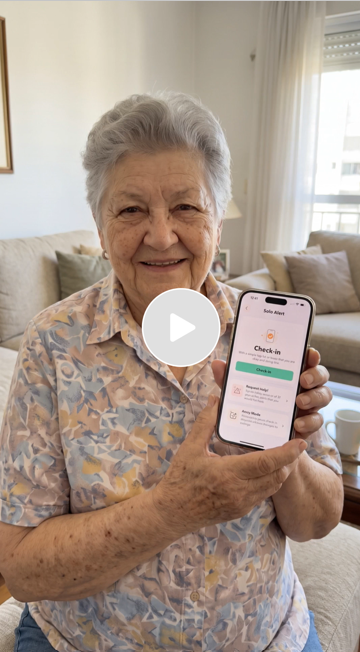 An elderly woman with gray hair smiling and holding a smartphone displaying a check-in screen. She is sitting in a bright living room with curtains and sofas in the background.