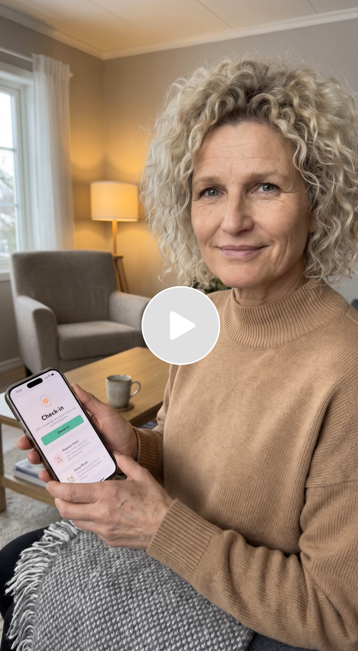 A woman with curly gray hair sitting on a couch, holding a smartphone displaying a health check app, in a living room with a lamp and armchair behind her.