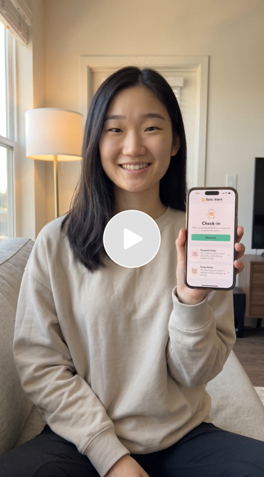 Young woman sitting on a couch holding a smartphone displaying a health app with a green checkmark and check-in reminder in a cozy living room with beige walls, a lamp, and a window.