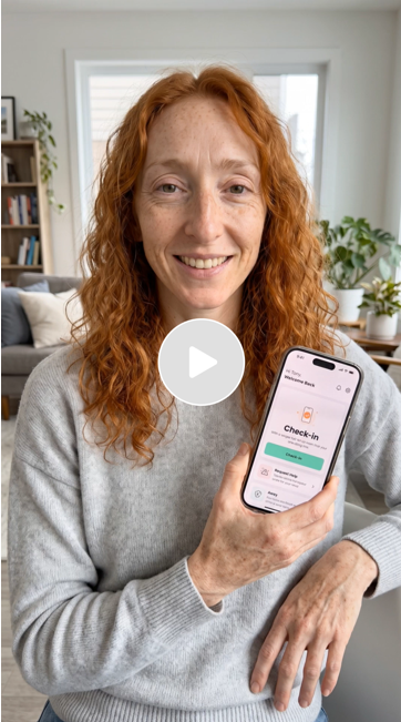 A woman with curly red hair holding a smartphone showing a check-in app, sitting in a bright living room with plants and shelves.