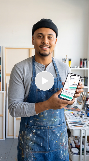 A man wearing a gray shirt, black beanie, and blue apron holding a smartphone with a check-in app, standing in an art studio with paint supplies and artwork in the background.