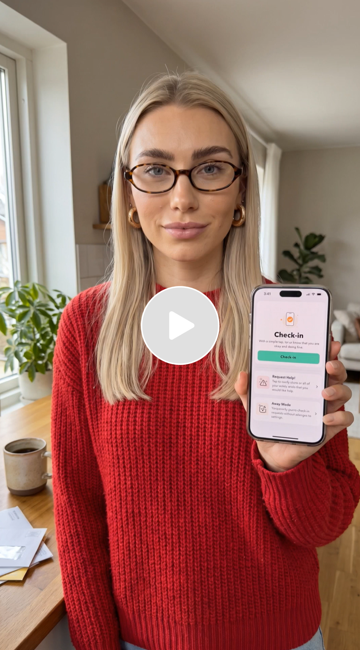 Woman with glasses and blonde hair wearing a red sweater, holding a smartphone displaying a check-in app, standing in a bright room with a window, a potted plant, and a desk with papers on it.