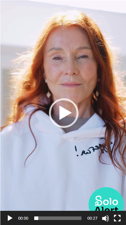 A woman with long red hair, smiling softly at the camera, outdoors in sunlight, wearing a white hoodie with a small logo and a turquoise circle with 'Solo Alert' printed on it.