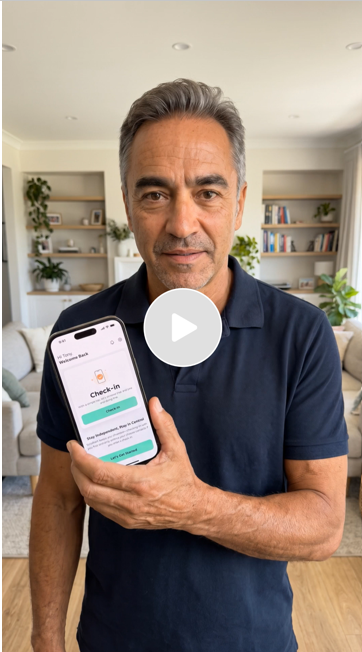 A middle-aged man with gray hair holding a smartphone displaying a check-in app, standing in a living room with shelves, plants, and a beige couch in the background.
