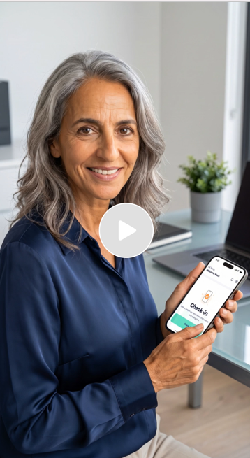 A smiling older woman with gray hair sitting at a desk, holding a smartphone displaying the Checkly app, in a bright office with a laptop and a potted plant.