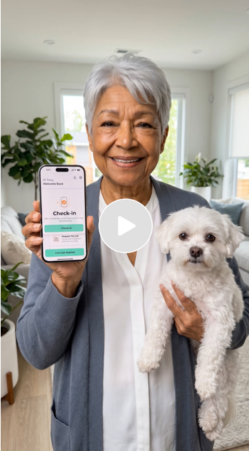 An elderly woman smiling while holding a dog and showing her phone with a check-in screen on it in a bright living room.