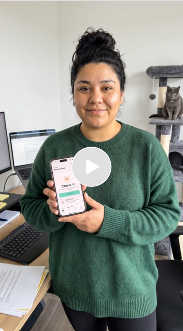A woman smiling and holding a smartphone displaying a check-in app in an office with a laptop, papers, and a gray cat on a multi-level cat tree in the background.