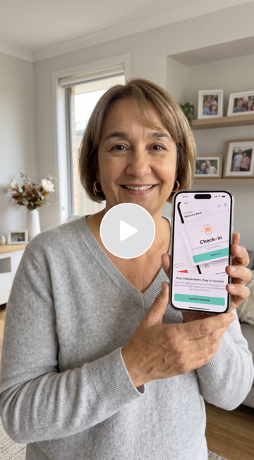 Smiling woman in gray sweater holding up smartphone with checking-in app open in living room with photo frames and flowers in background.