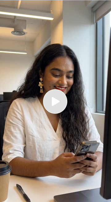 A woman with long curly hair, wearing a white blouse and gold earrings, is sitting at a desk looking at her phone and smiling.